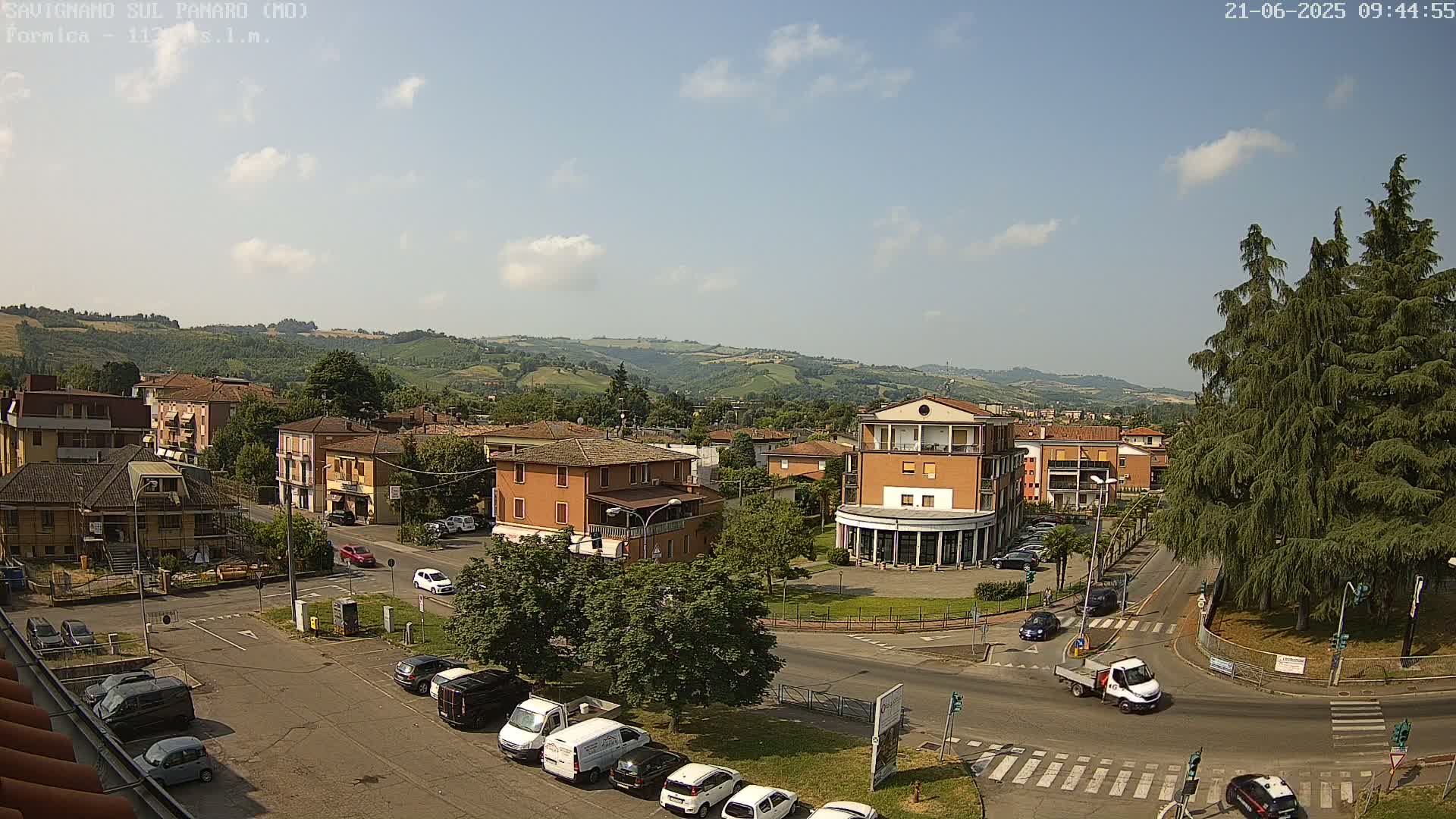 A sunny day reveals a view of a town square surrounded by low buildings, parked cars, and a few passing vehicles, all nestled within a hilly landscape.
