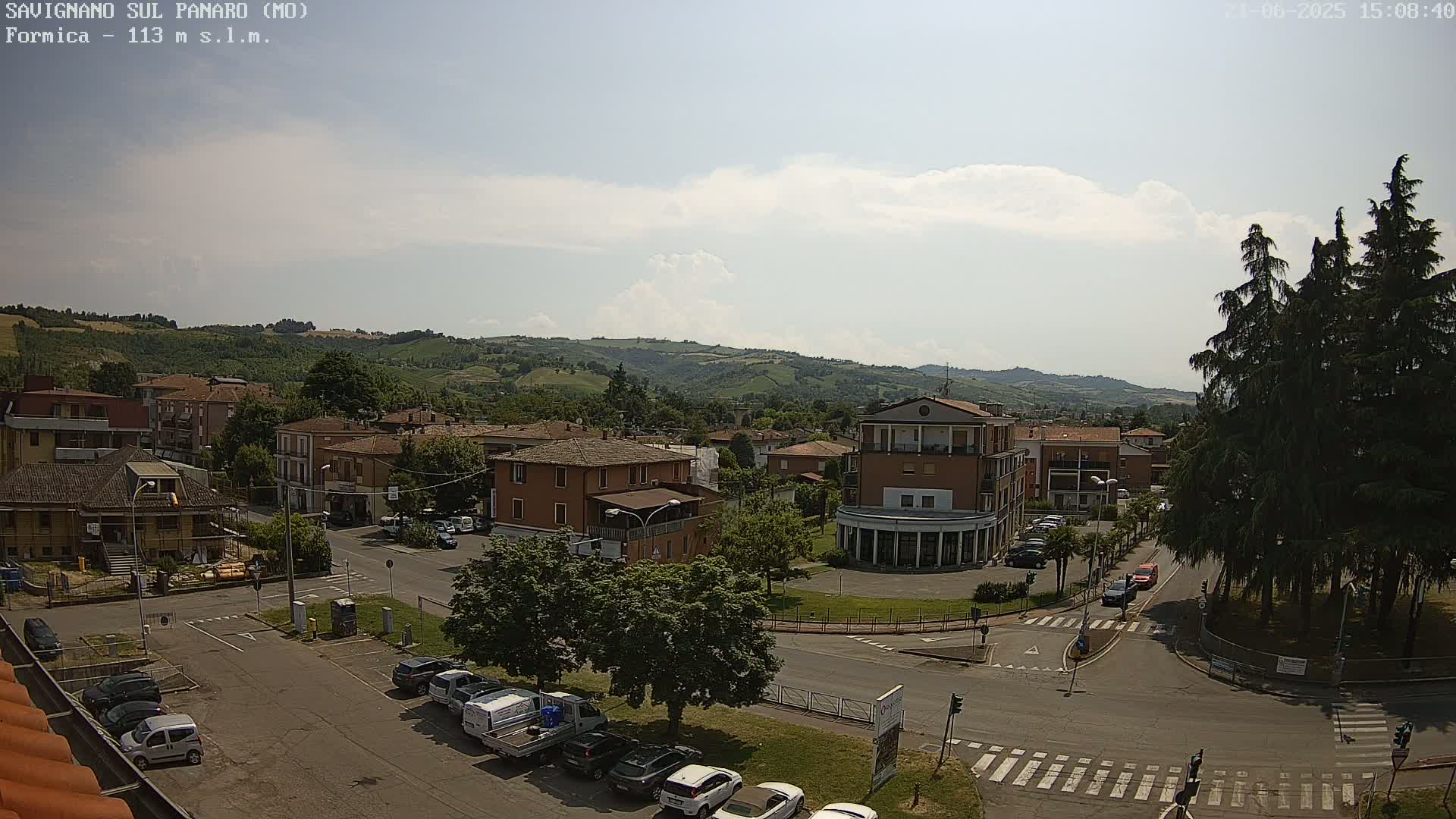 A partly cloudy sunny day overlooks a town nestled in a valley, surrounded by green hills and several cars parked along roadways.