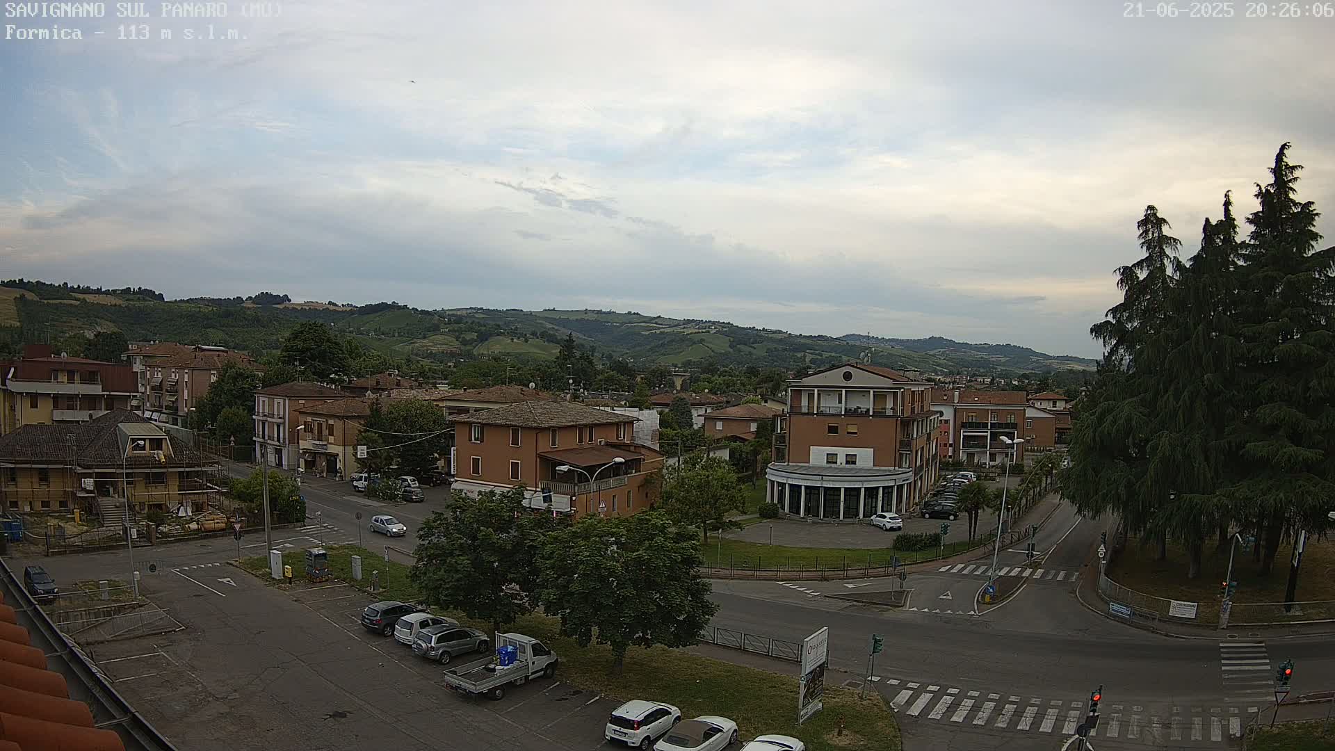 A partly cloudy day reveals a view of a town nestled in a valley, with cars parked in a lot and buildings surrounding a small plaza.