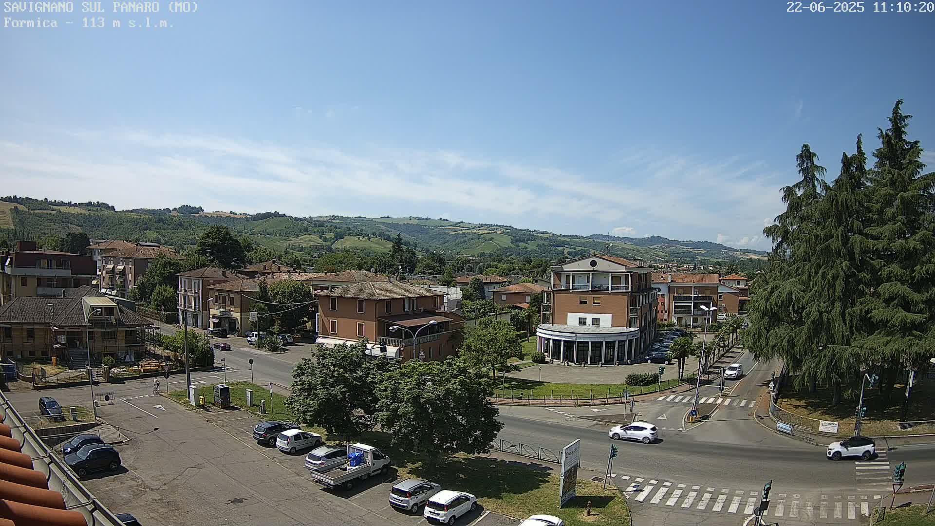 A sunny day reveals a town nestled in a valley, with cars parked along streets and several buildings visible, surrounded by green hills.