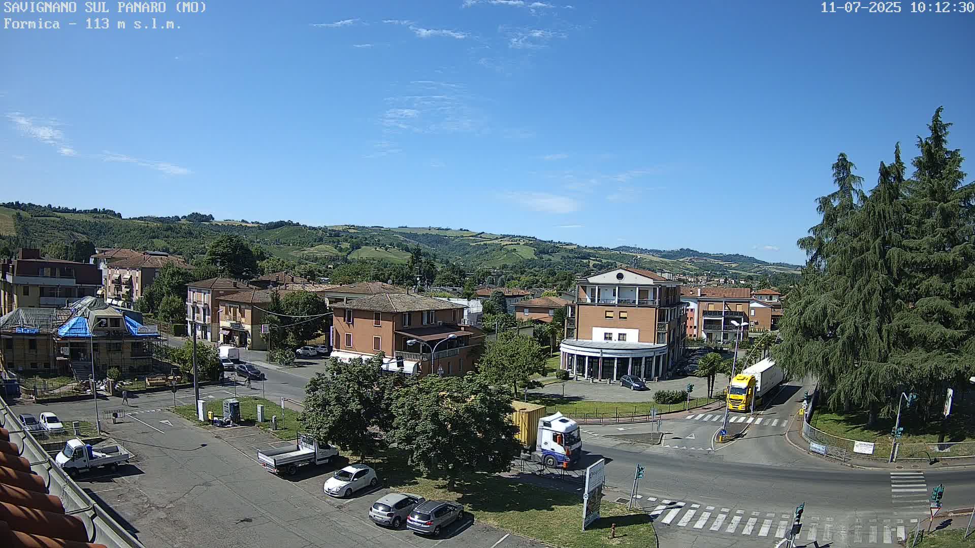 A sunny day reveals a town square with buildings, vehicles, and trees, nestled in a valley with rolling green hills in the background.