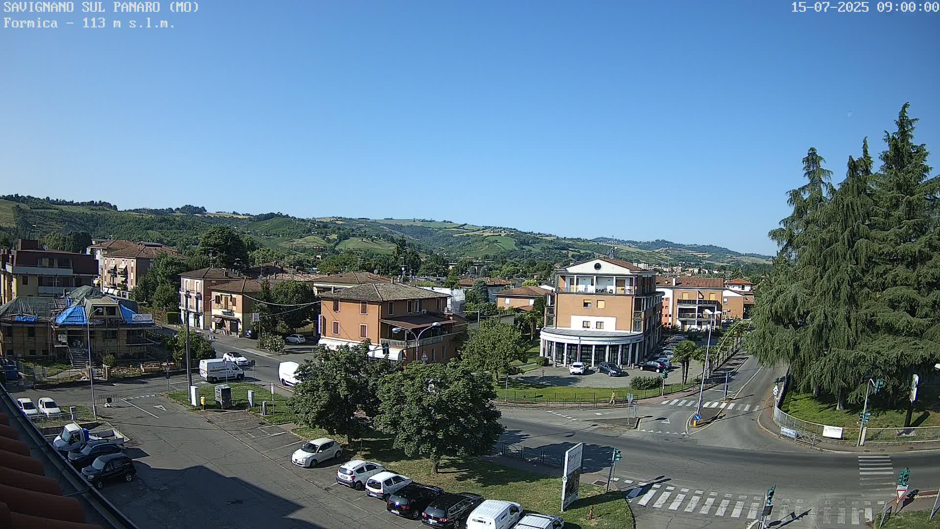 A sunny, clear-day view of a town square in a small Italian town, nestled in a valley surrounded by rolling green hills, shows several buildings, parked cars, and some trees.