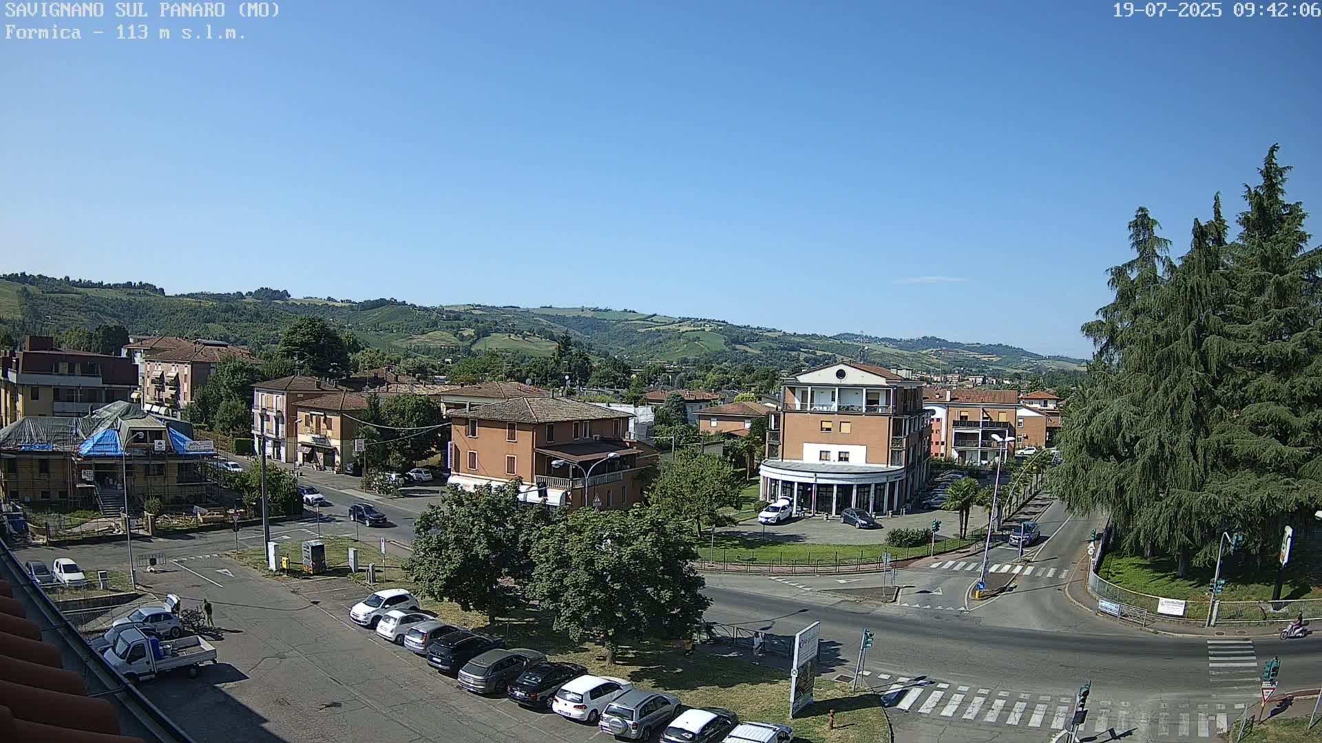 A sunny, high-angle view of a town square with buildings, cars, and a large evergreen tree, set against a backdrop of rolling hills under a clear blue sky.