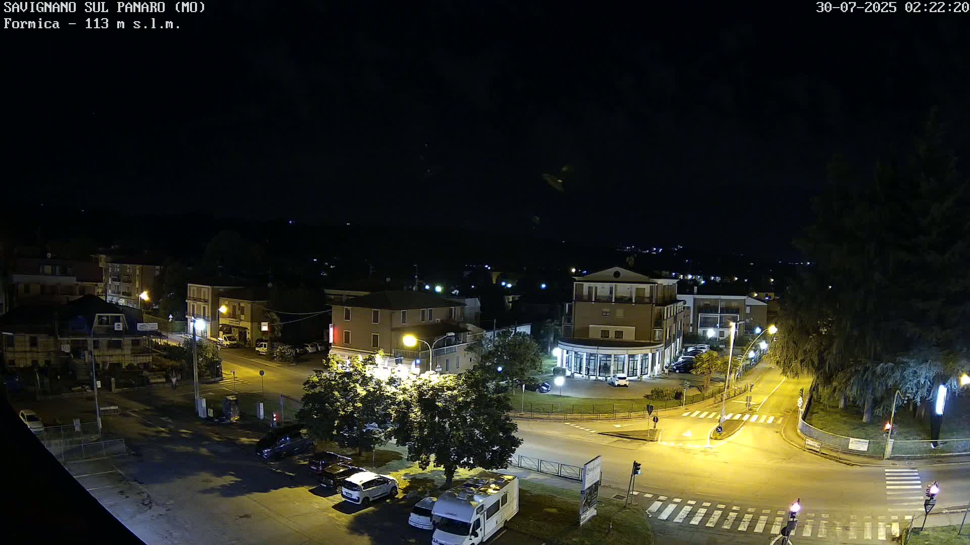 A sunny, high-angle view of a town square with buildings, cars, and a large evergreen tree, set against a backdrop of rolling hills under a clear blue sky.