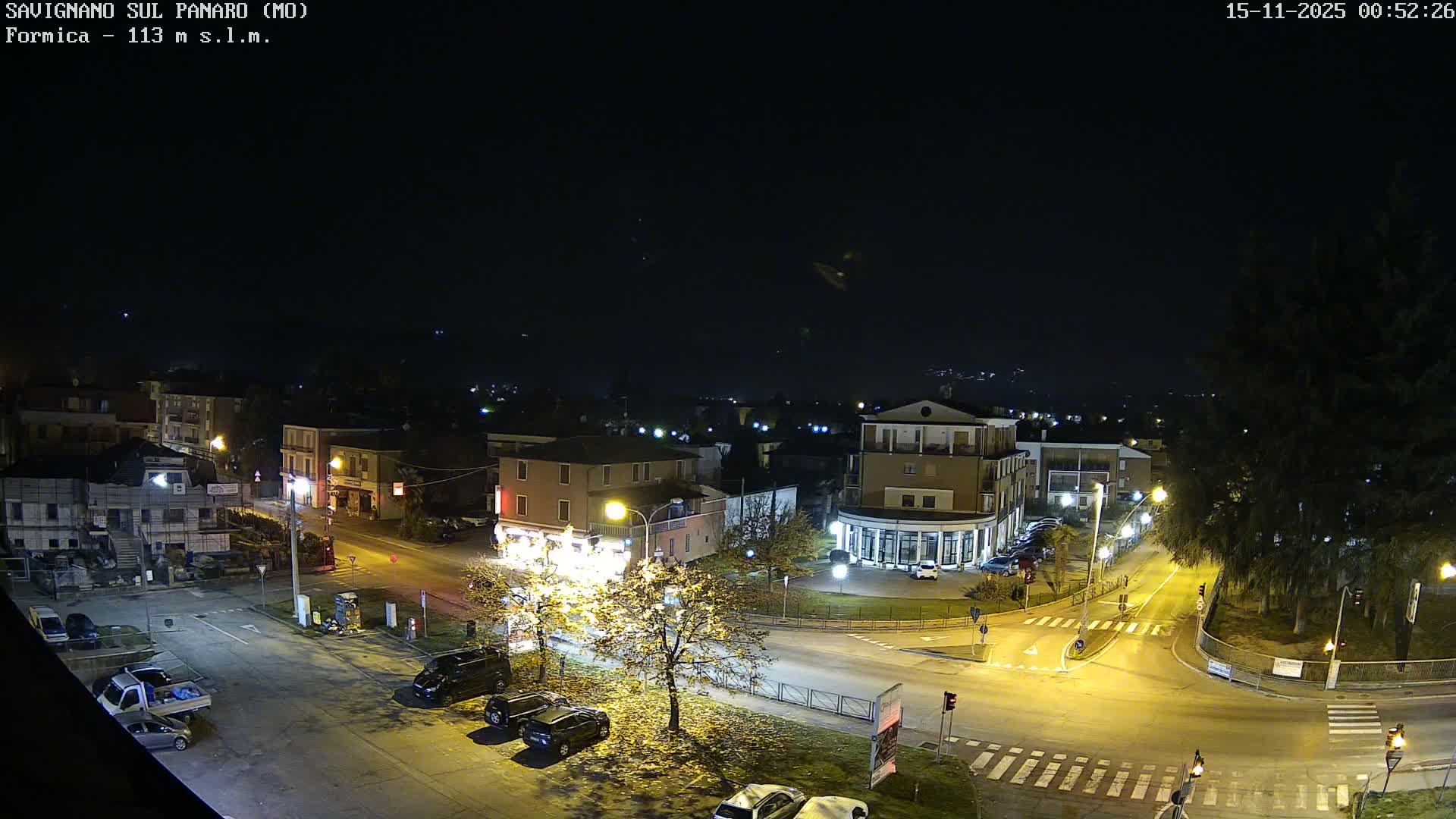An aerial nighttime view shows a brightly lit town intersection with buildings, parked cars, and trees, all under a clear dark sky.