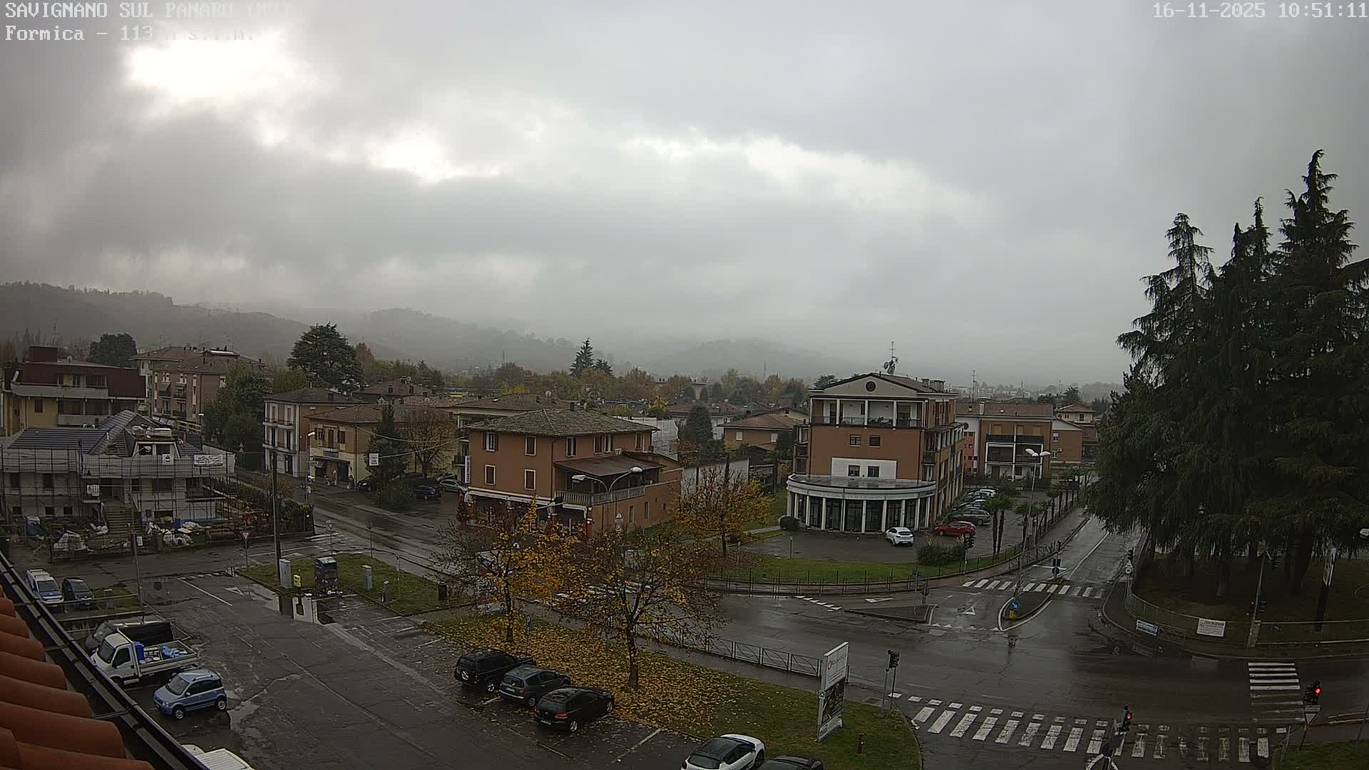 An elevated view shows a town under an overcast sky with wet roads, featuring various buildings, parked cars, and trees with autumn foliage, all set against a backdrop of misty hills.