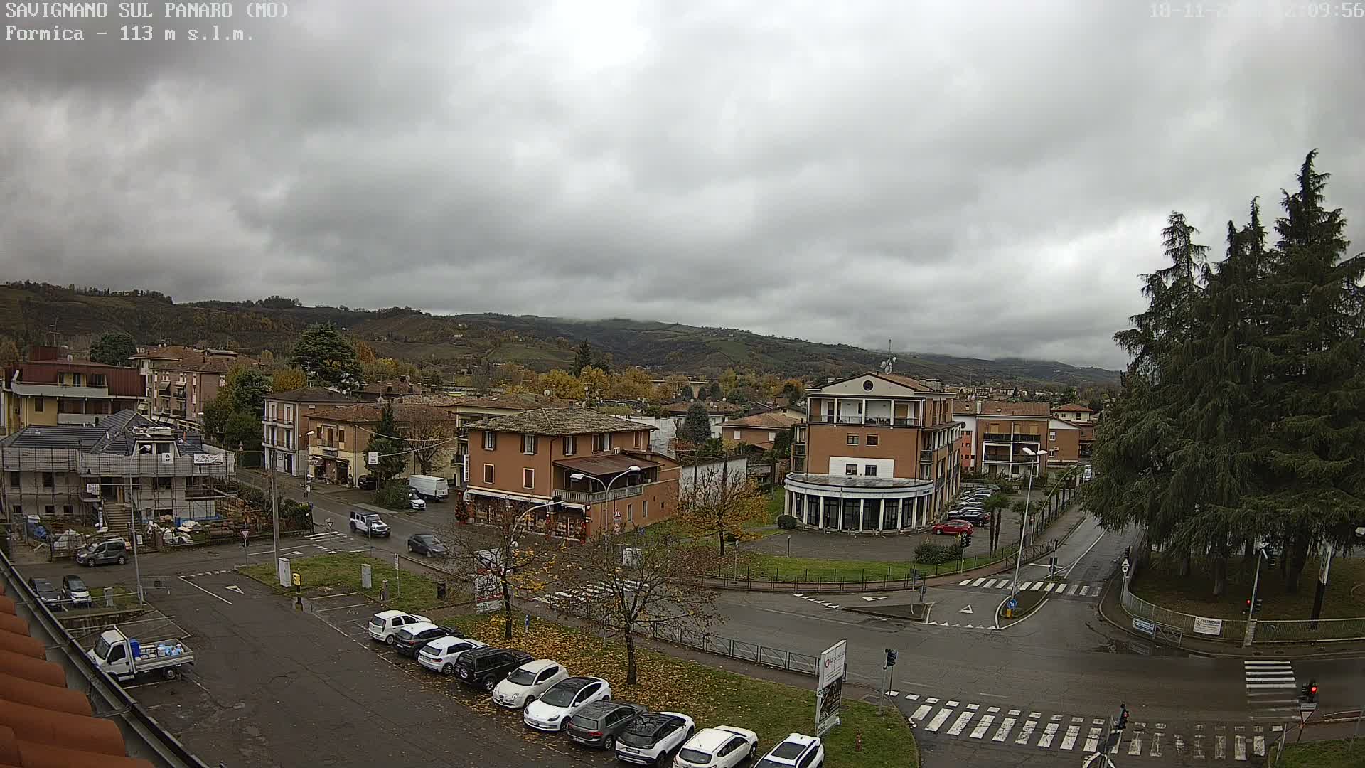 An elevated view shows a town under an overcast sky with wet roads, featuring various buildings, parked cars, and trees with autumn foliage, all set against a backdrop of misty hills.