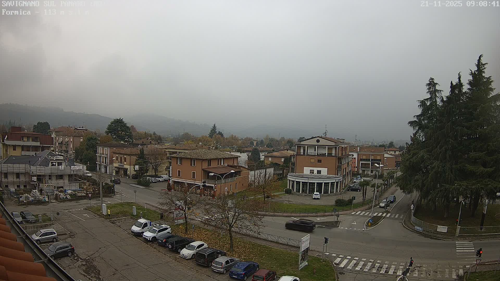 A town with a mix of buildings, roads, and cars, including one under construction, stretches towards hazy, tree-covered hills under a uniformly overcast and foggy sky.