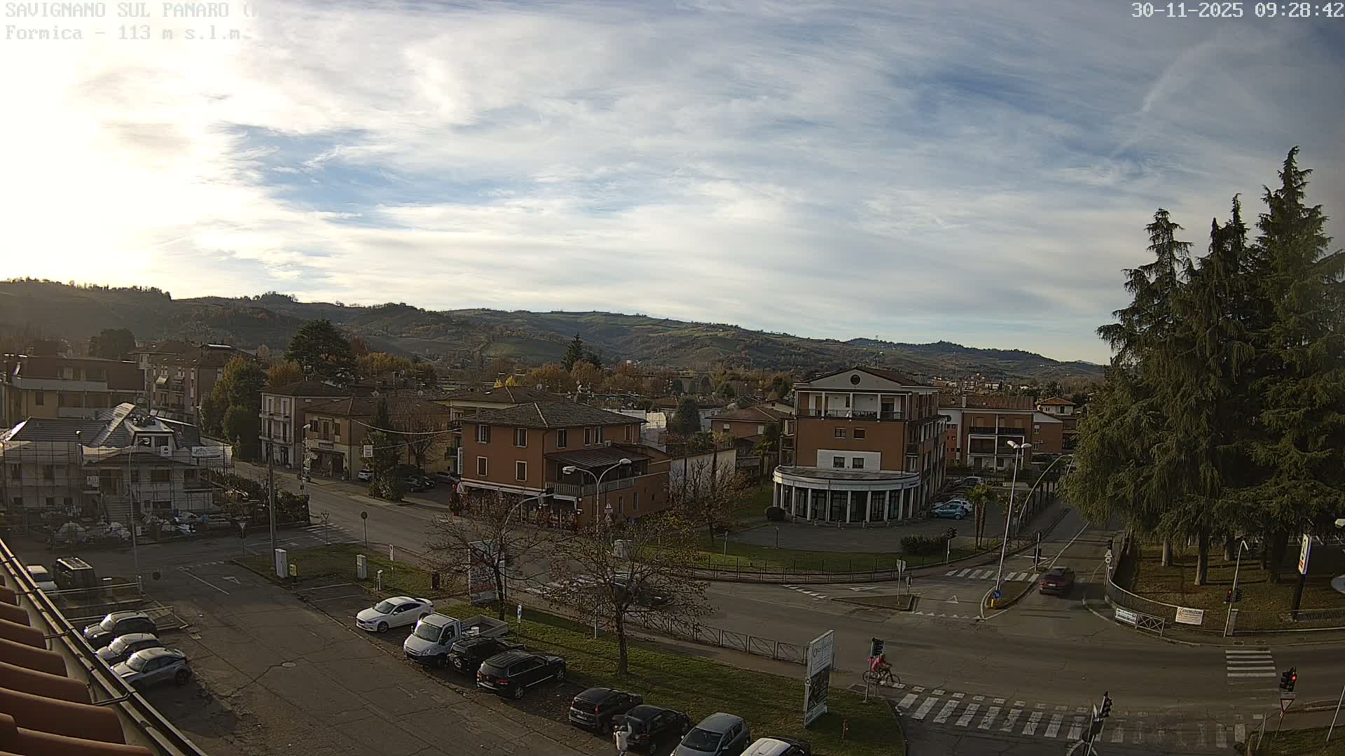 The image shows an elevated view of a town with various buildings and streets, including a busy intersection with parked cars and a few vehicles, all set against a backdrop of rolling, tree-covered hills under a partly cloudy sky with patches of blue.