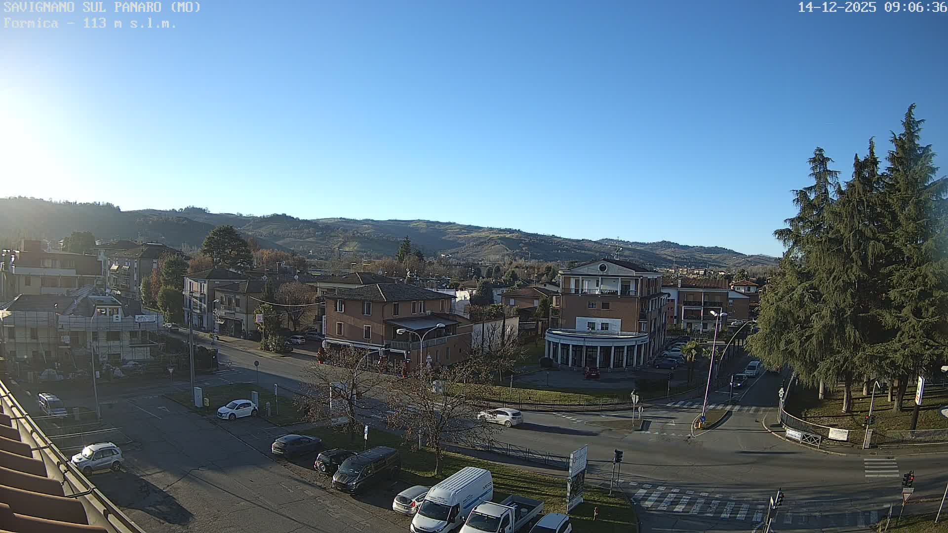 This image captures a clear, sunny day over a European town featuring buildings and streets with parked cars, nestled against a backdrop of rolling, partially vegetated hills under a bright blue sky.