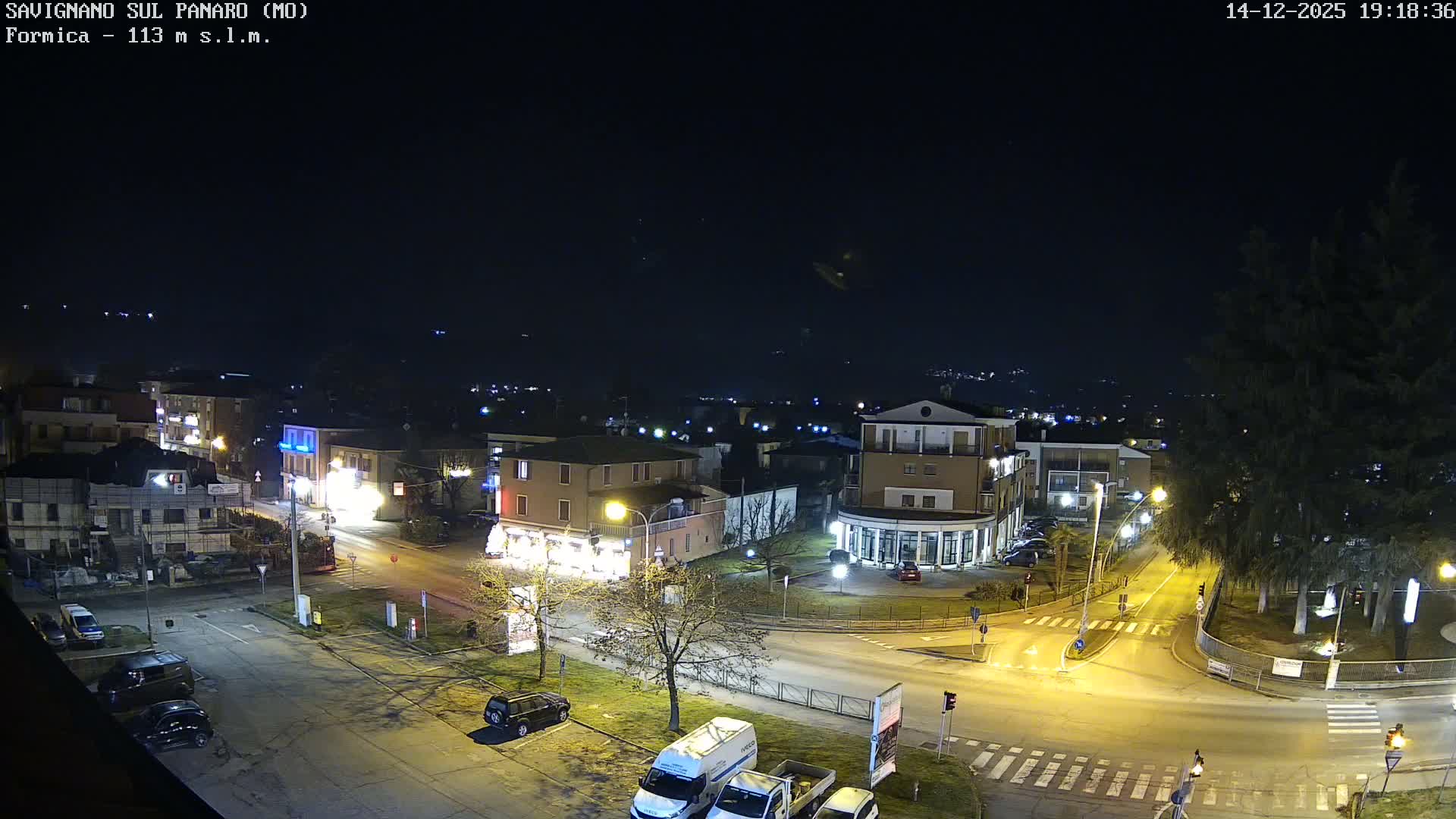 This image captures a clear, sunny day over a European town featuring buildings and streets with parked cars, nestled against a backdrop of rolling, partially vegetated hills under a bright blue sky.