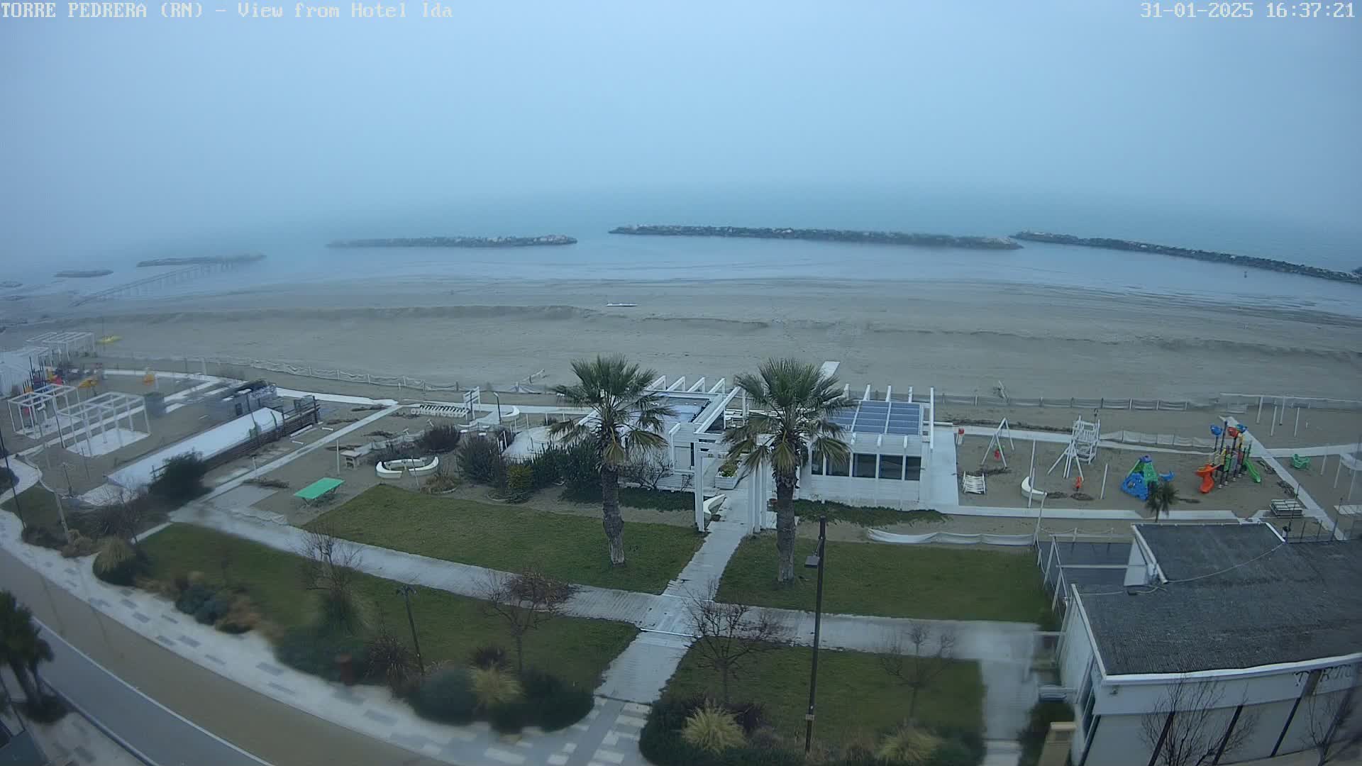 A hazy, overcast day reveals a beach with a playground, a white building, palm trees, and a paved walkway leading to the ocean.