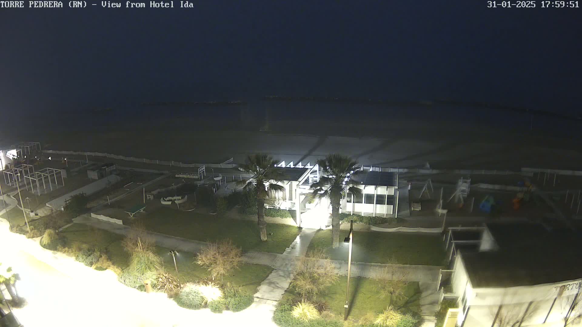 A nighttime, high-angle view shows a beach, beachfront buildings, and landscaping, all illuminated under a dark sky.