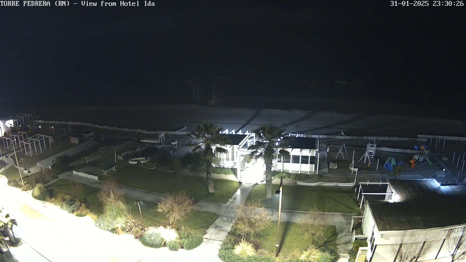A nighttime aerial view of a beach area shows a playground, some buildings, and palm trees, with a dark, seemingly calm sea in the background.