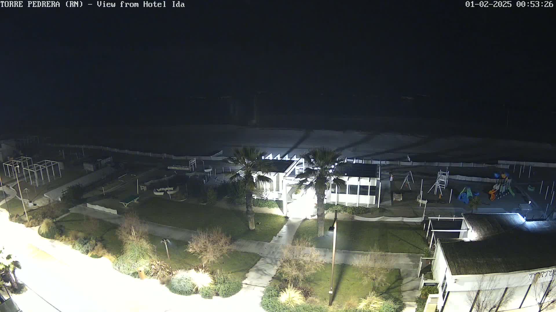 A nighttime aerial view shows a beach, a playground, and several buildings with palm trees, all illuminated under dark skies.