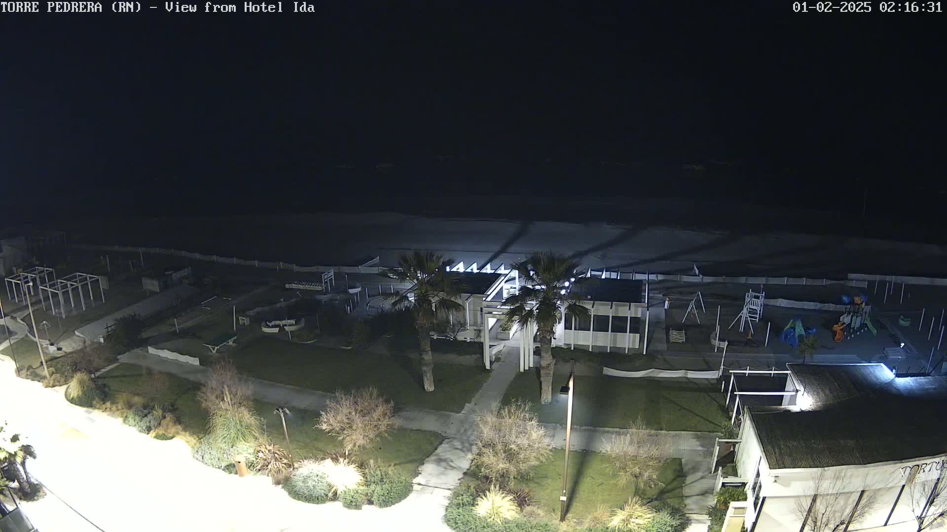 A nighttime aerial view shows a beach with a playground, some buildings, and palm trees, under a dark sky.