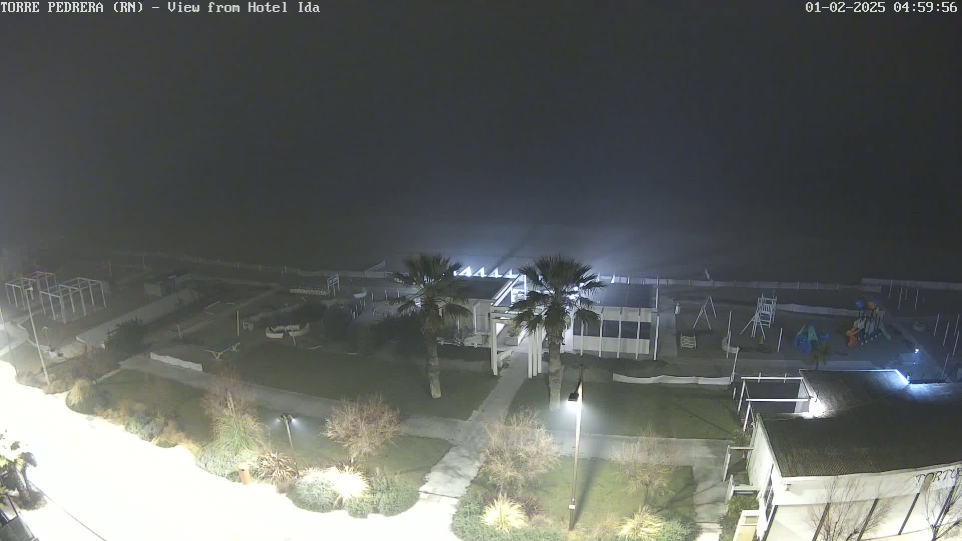 A nighttime, foggy view of a beachfront area shows a building with palm trees in front, a playground, and other outdoor recreational equipment.