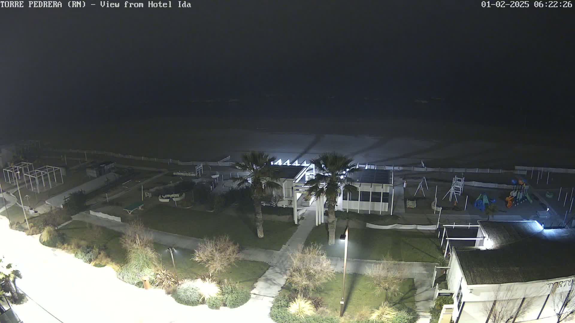 A nighttime view of a beach, playground, and building complex, with palm trees and landscaping visible under a dark sky.