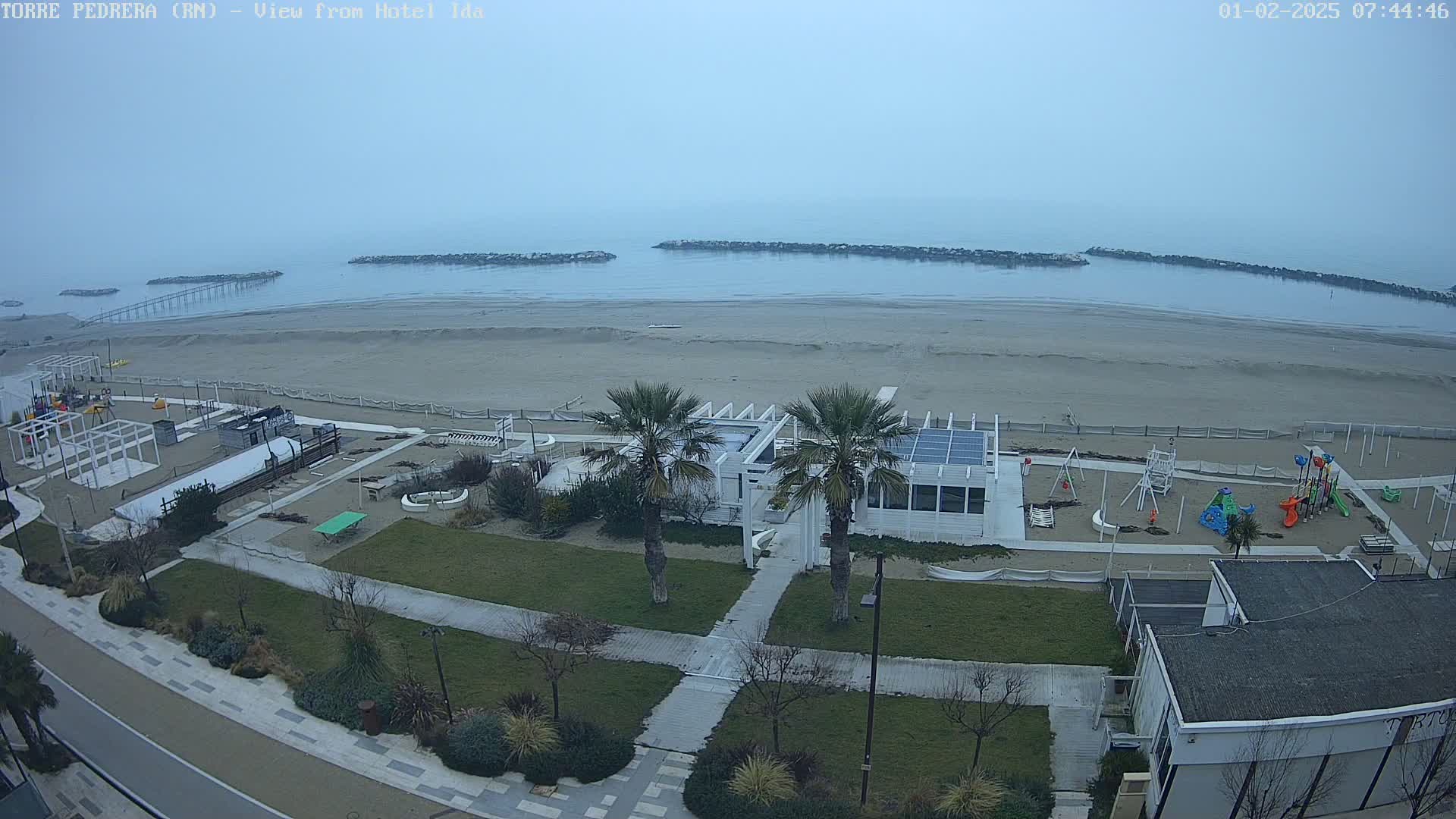 A beach scene on an overcast day shows a sandy beach, calm water, a playground, some palm trees, and a white building near the water.