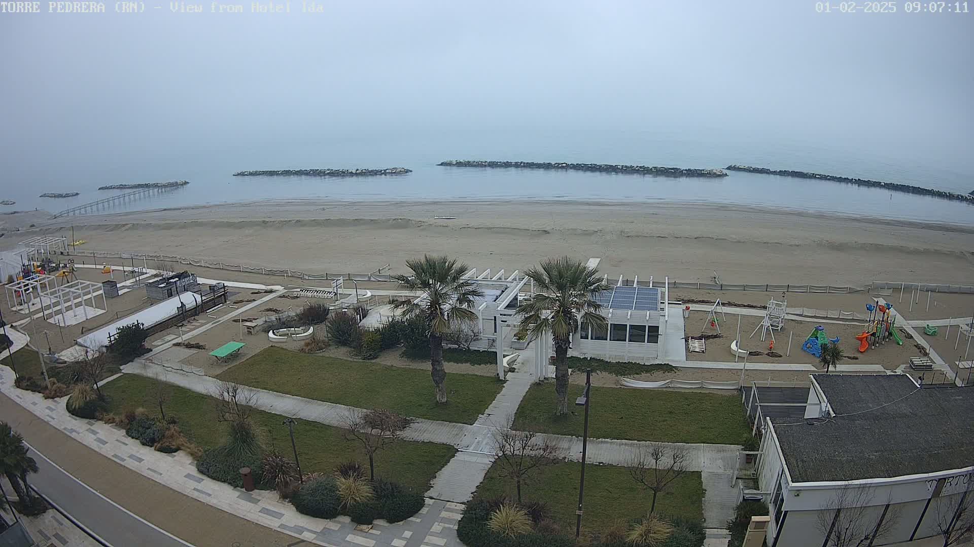 A beach with a playground, palm trees, and buildings is seen on an overcast day.