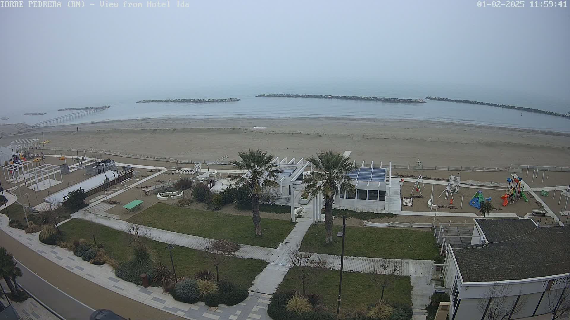 A beach with a playground, a white building, and palm trees is visible in the foreground, while a calm, foggy sea stretches out to the horizon.