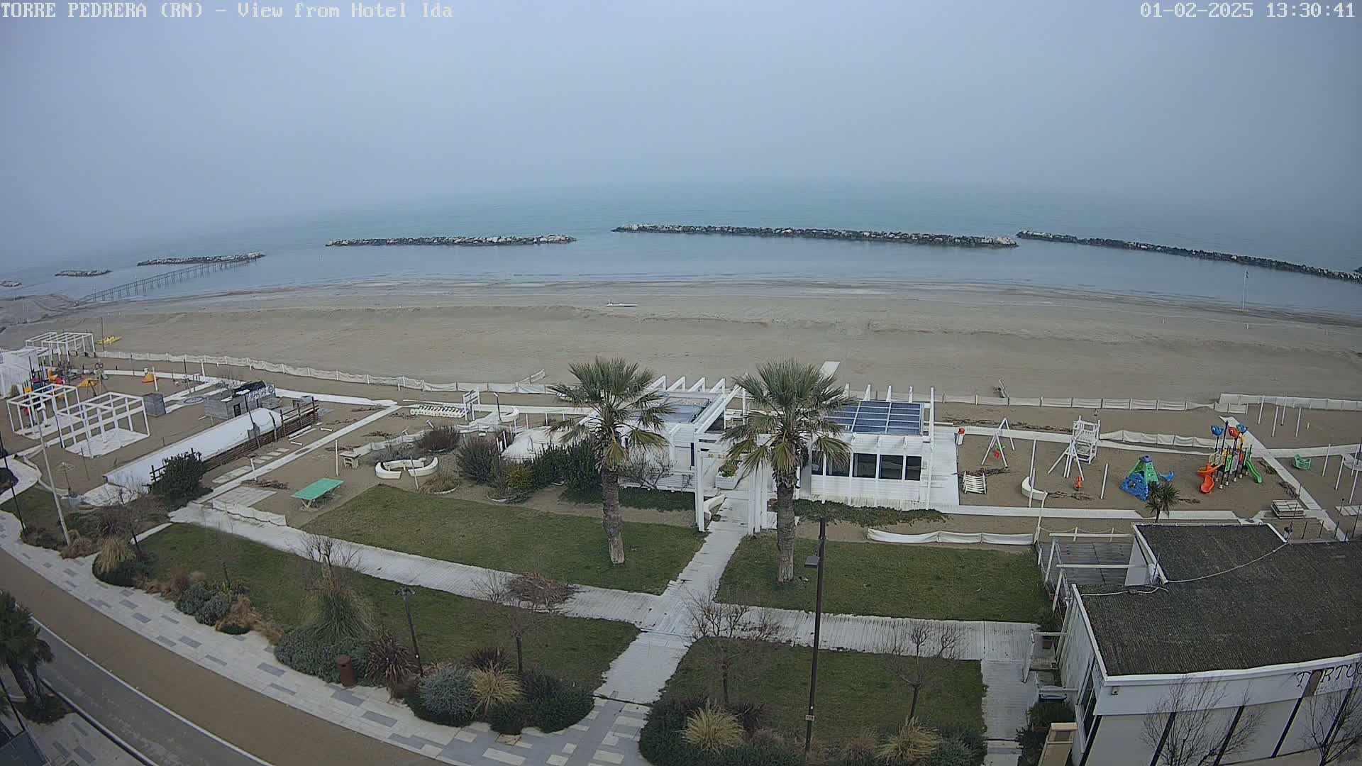 A beach with a playground and a white building is seen on an overcast day, with calm water extending to the horizon.