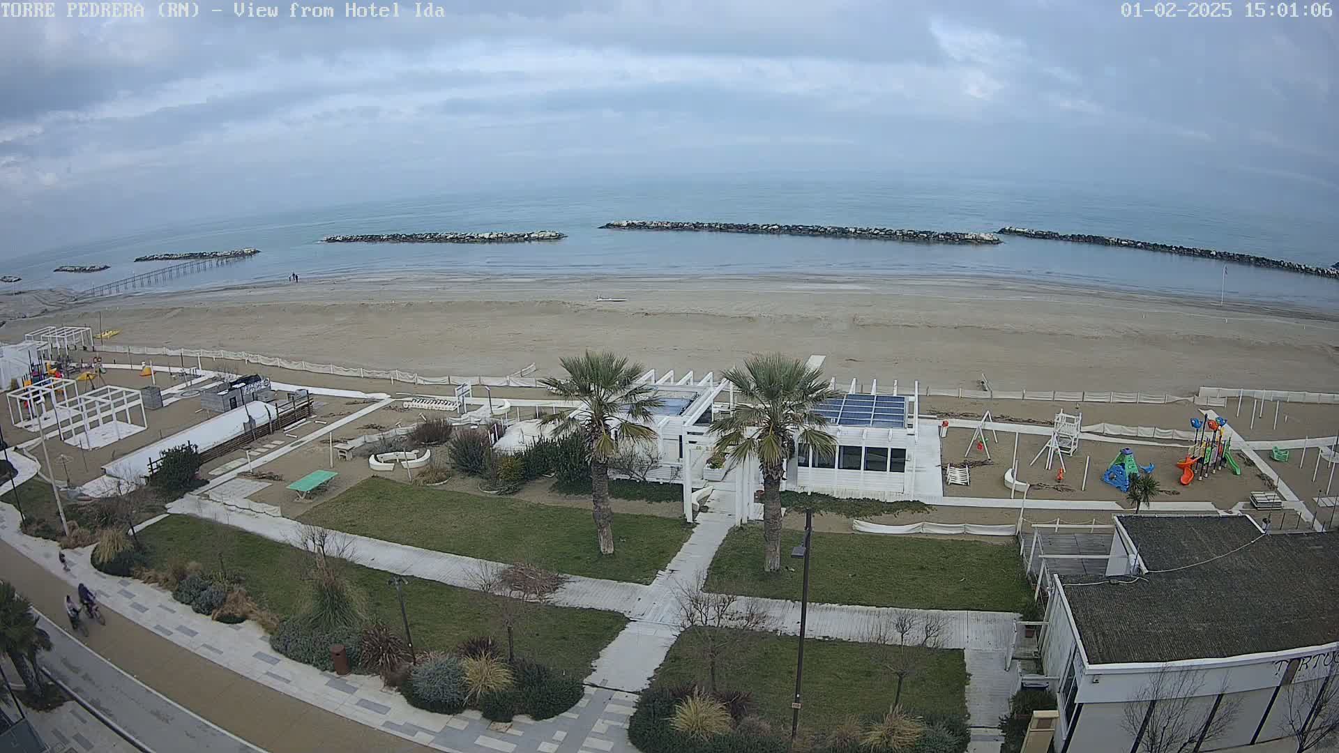 A beach with calm, overcast skies, bordered by a boardwalk with a playground and a white building, is viewed from above.