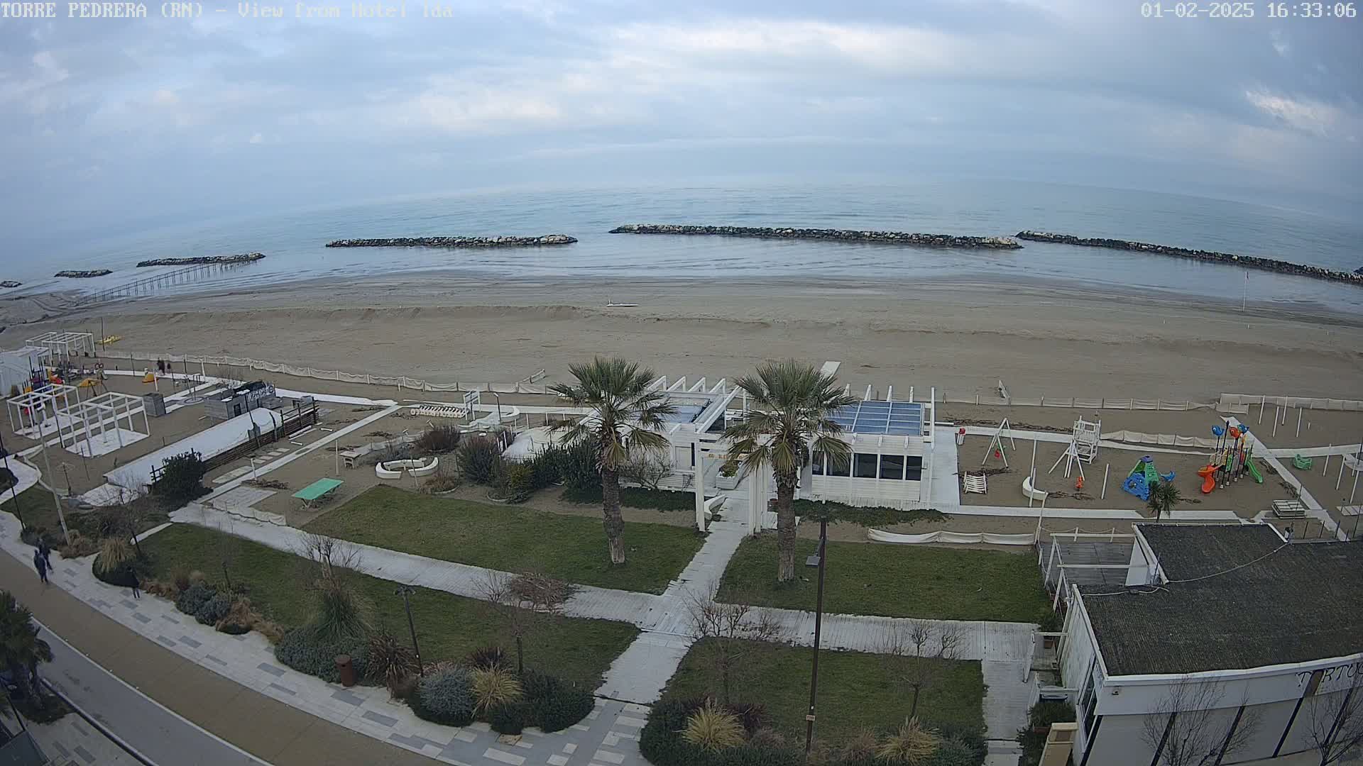 An overcast day at a beach shows a sandy shoreline with calm water, rock barriers, a playground, beachside buildings, and palm trees.