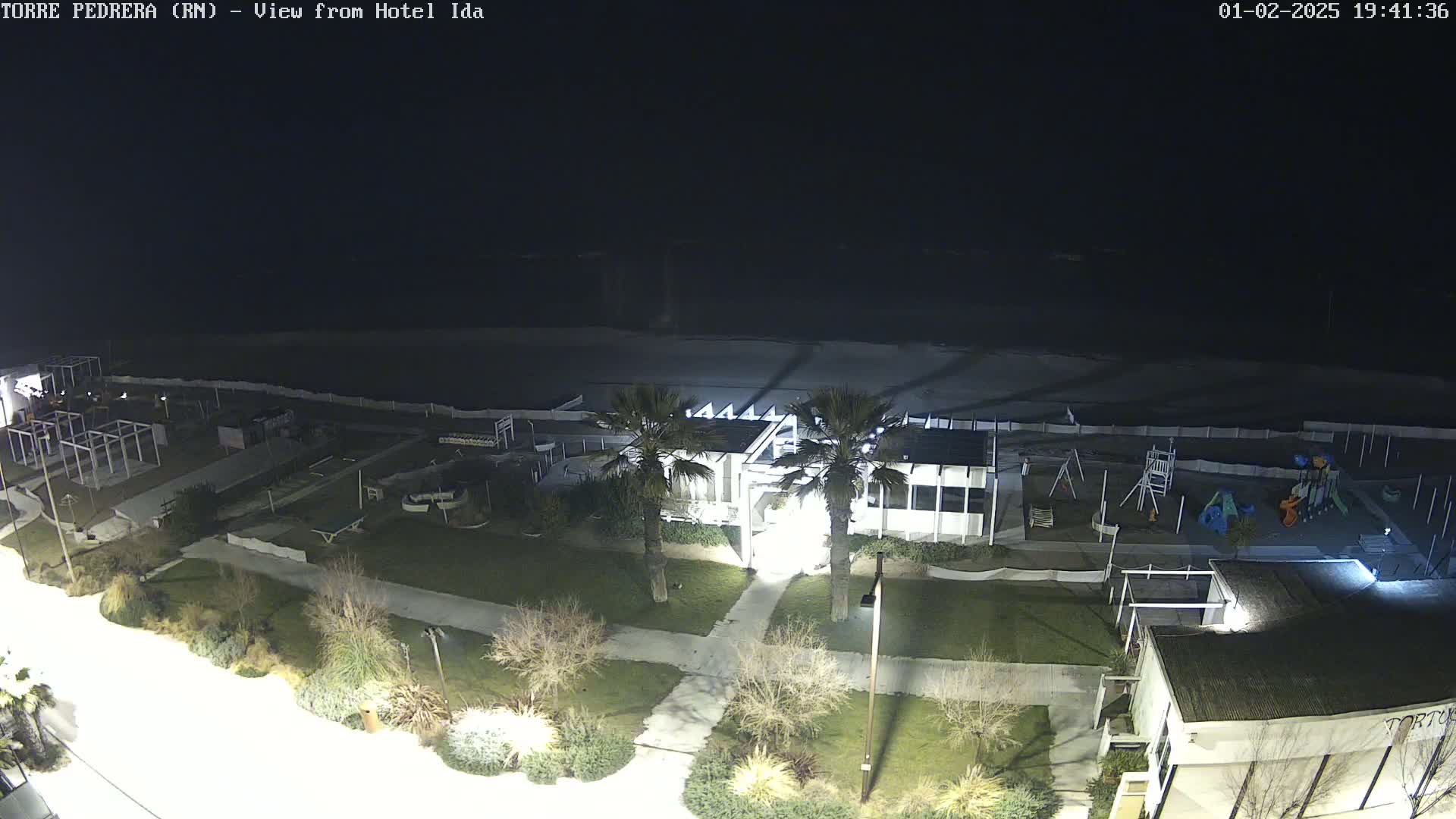 A nighttime aerial view of a beach area shows a building, playground, and sparsely populated grassy areas next to a dark ocean under a clear, dark sky.