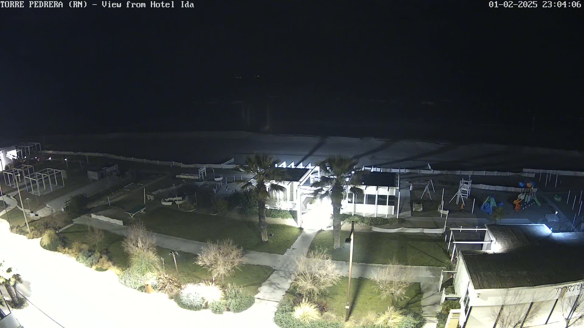 A nighttime aerial view of a beachside area shows a building, playground, and beach with calm water under a dark sky.