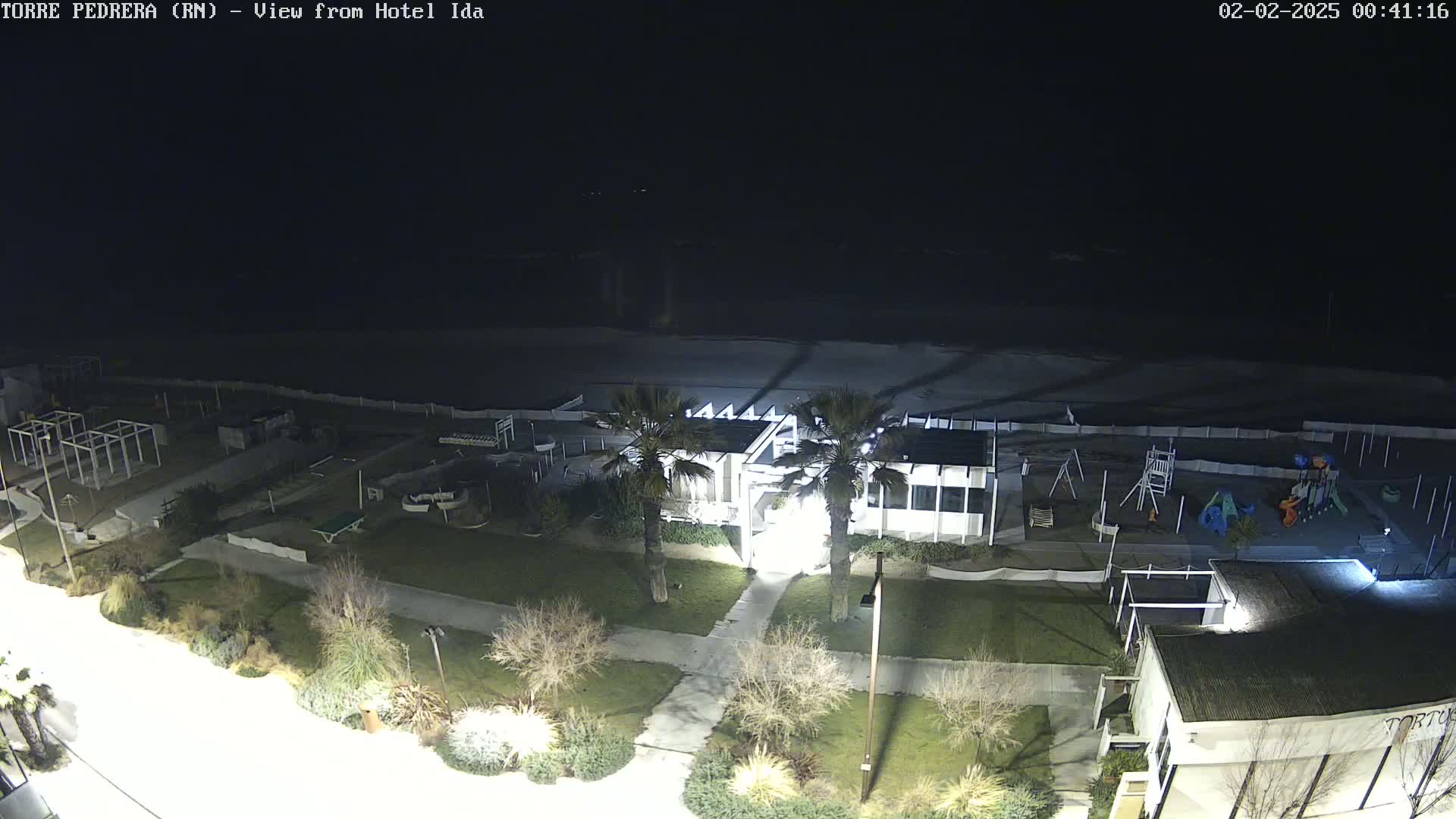 A nighttime view of a beachfront area shows a building, playground, and beach with calm water under a dark sky.