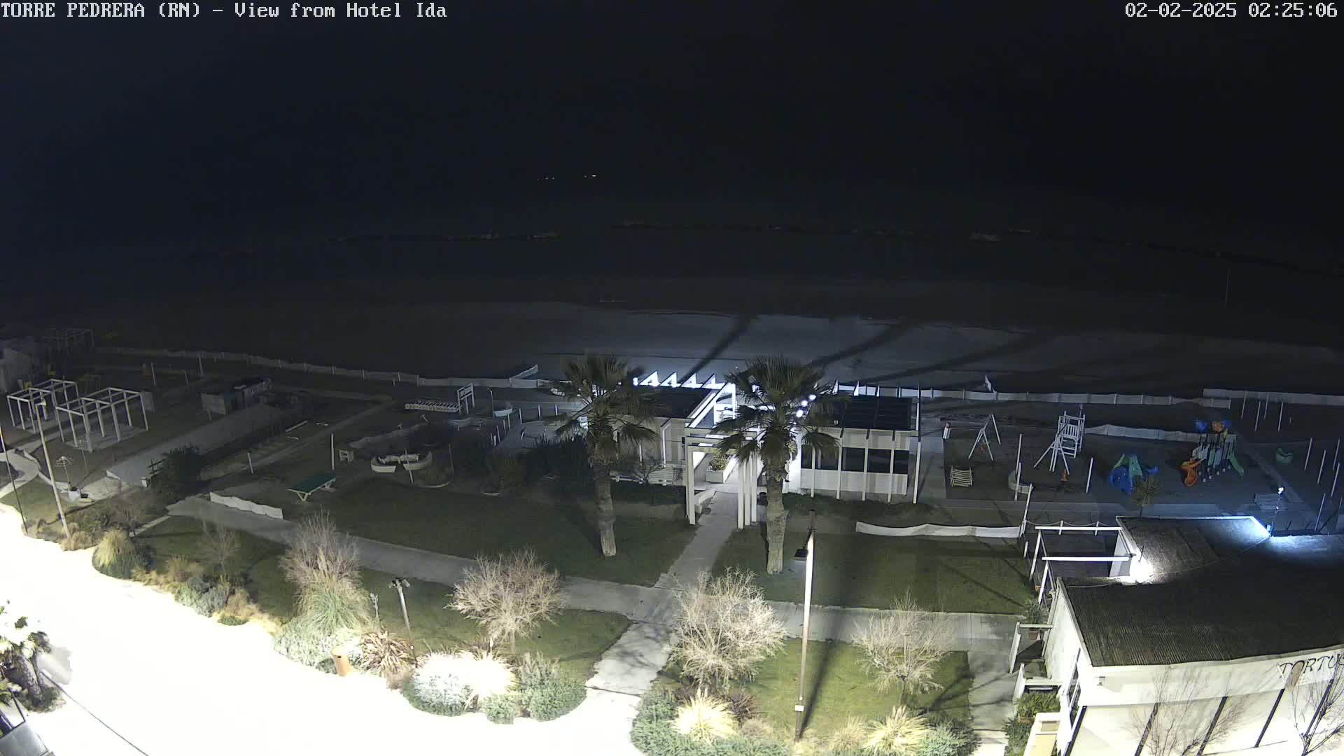 A nighttime aerial view of a beach, playground, and beachfront building illuminated by artificial lights, with a dark, calm ocean in the background.