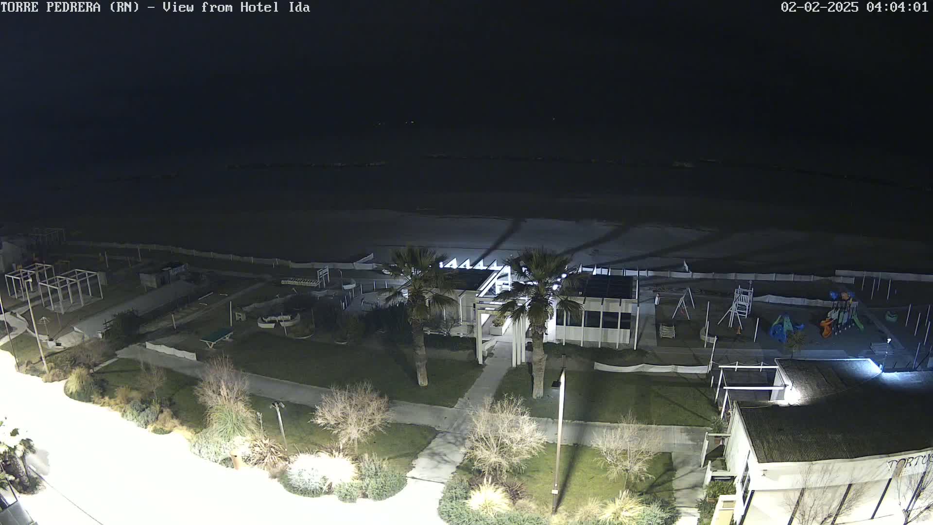 A nighttime aerial view shows a beach, a playground, a building, palm trees, and a roadway, under a clear, dark sky.