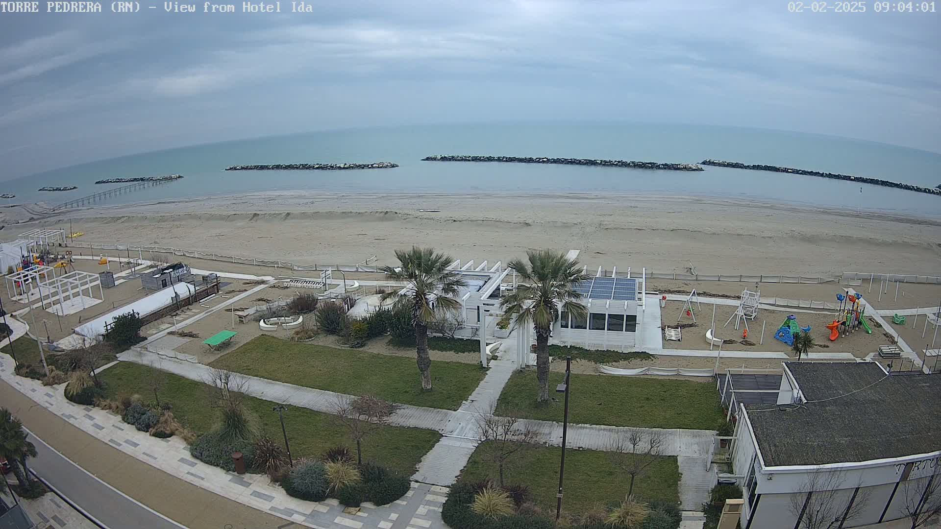 An overcast day reveals a sandy beach with a playground, beachfront restaurant, and walkways, all situated near a calm ocean with breakwaters.