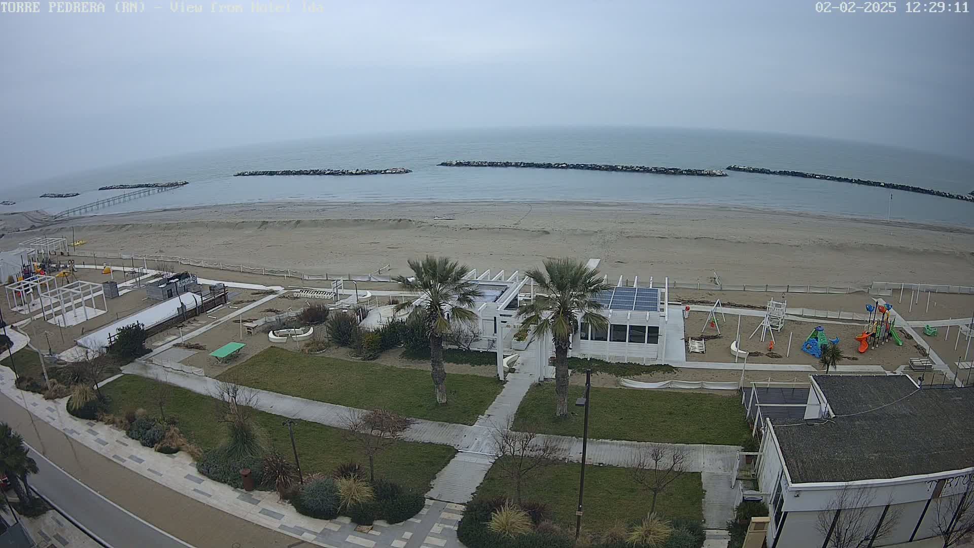 A sandy beach with calm, grey water under an overcast sky is shown, next to a playground and a white building with palm trees in front.