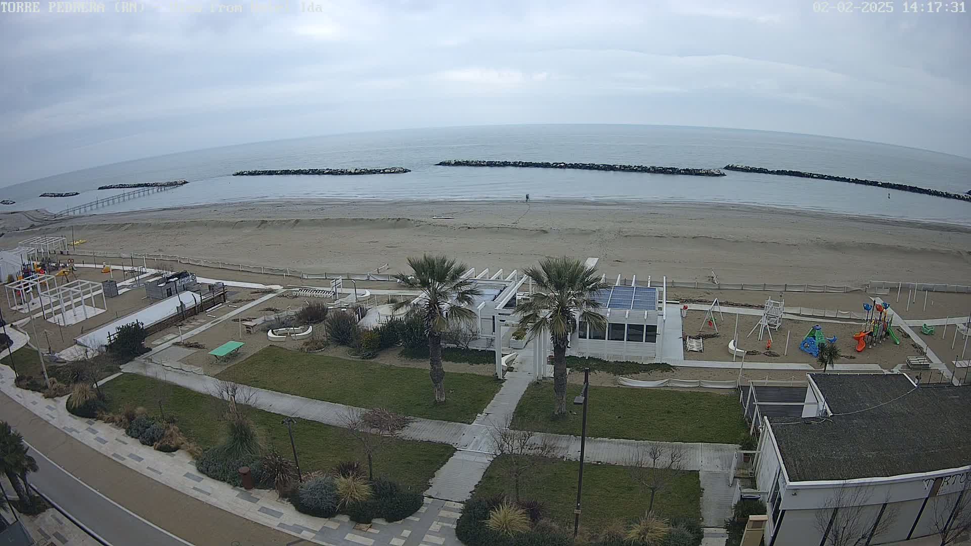 An overcast day reveals a sandy beach with a playground, beachfront building, and palm trees, all adjacent to a paved walkway and road.