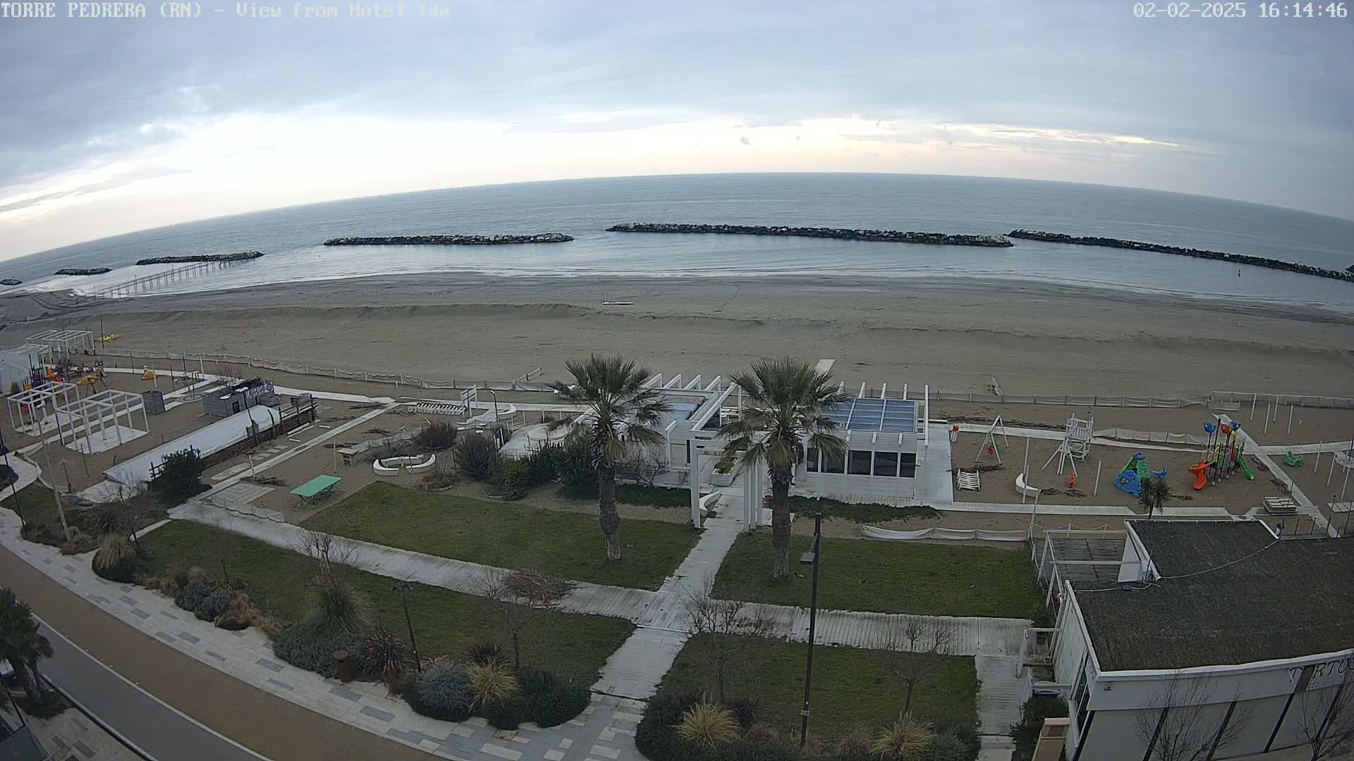 An overcast day reveals a sandy beach with calm ocean waves, rock barriers, a playground, and a white building with palm trees near a paved walkway.