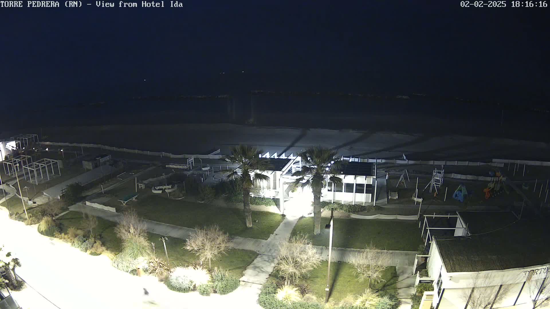 A nighttime aerial view of a beach area with a building, palm trees, and playground equipment, under a dark sky.