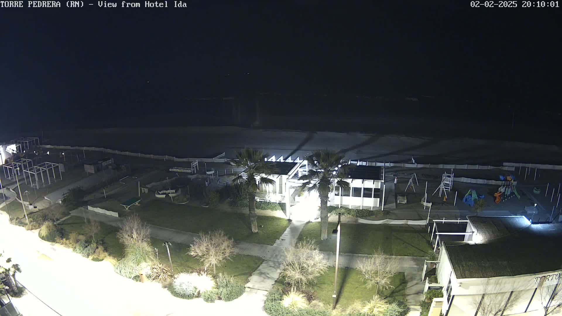 A nighttime, overhead view of a beachside area shows a building, playground, and palm trees under a dark sky.