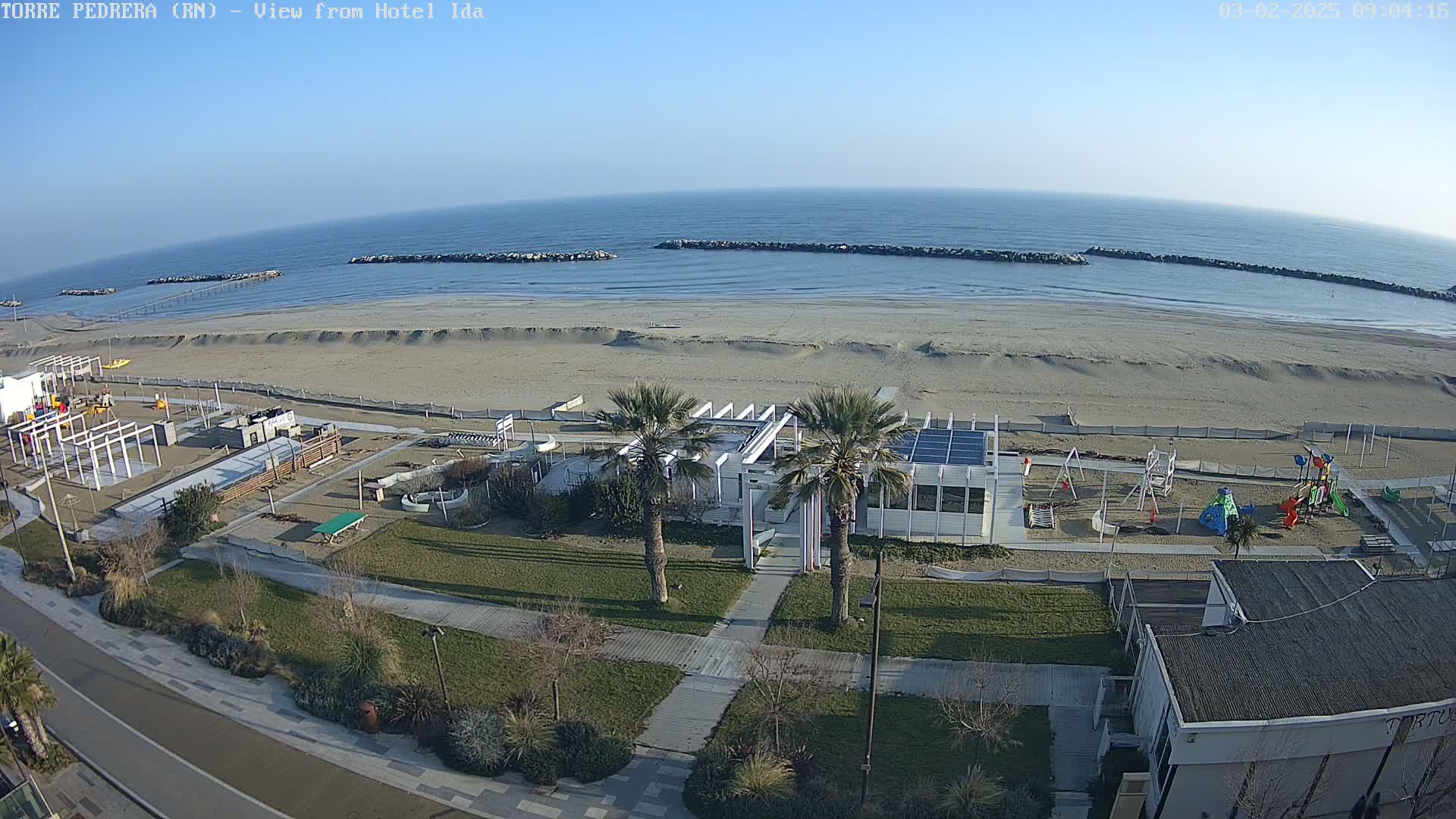 An aerial view shows a beach with calm ocean waves under a clear sky, a playground, a building with solar panels, and palm trees bordering a paved walkway.