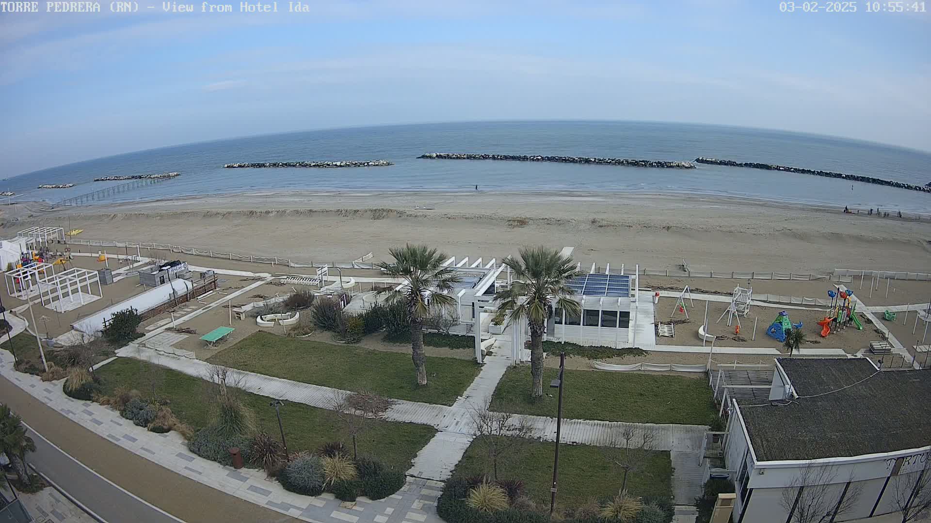 An aerial view shows a sandy beach with calm ocean water under a mostly clear sky, adjacent to a beachfront area with a playground, palm trees, and a white building.