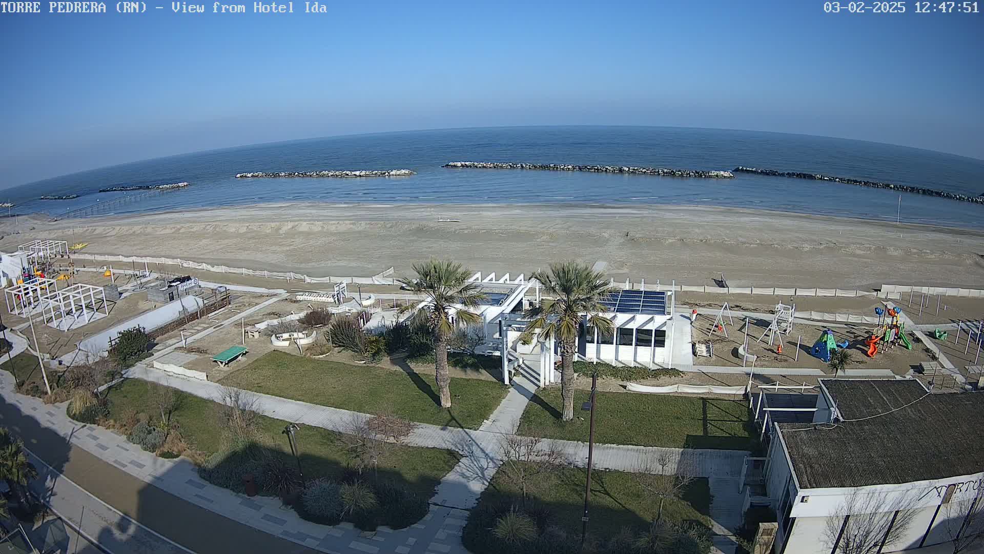 A beachfront area with a playground, a modern white building, and palm trees, all under a sunny sky.