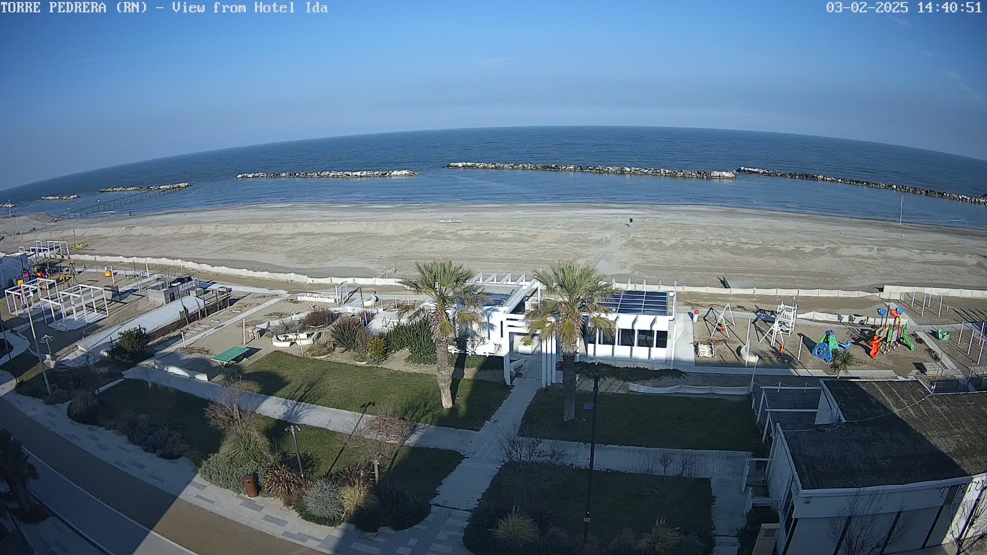 A sandy beach with calm ocean water under a clear sky, features a playground,  palm trees, and a white building near the shoreline.