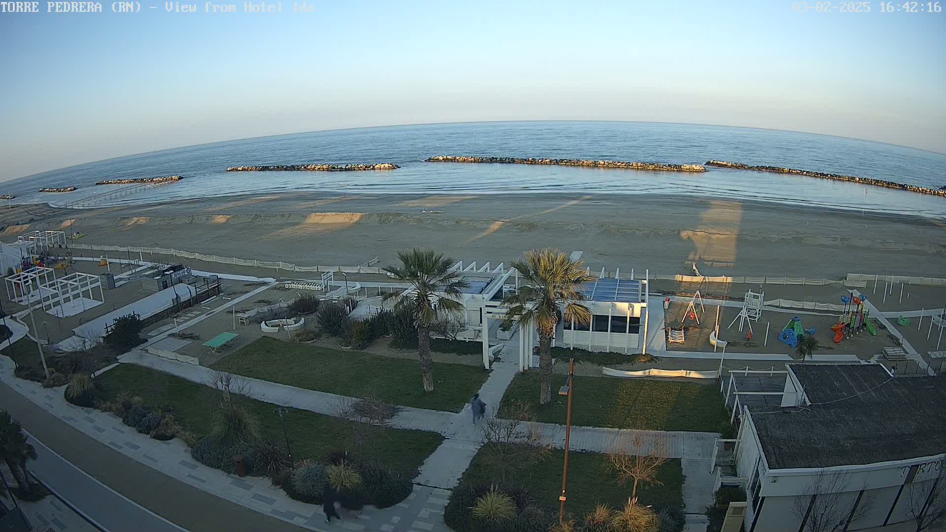 A sandy beach with calm ocean water, breakwaters, a playground, palm trees, and a white building is visible on a clear, sunny day.