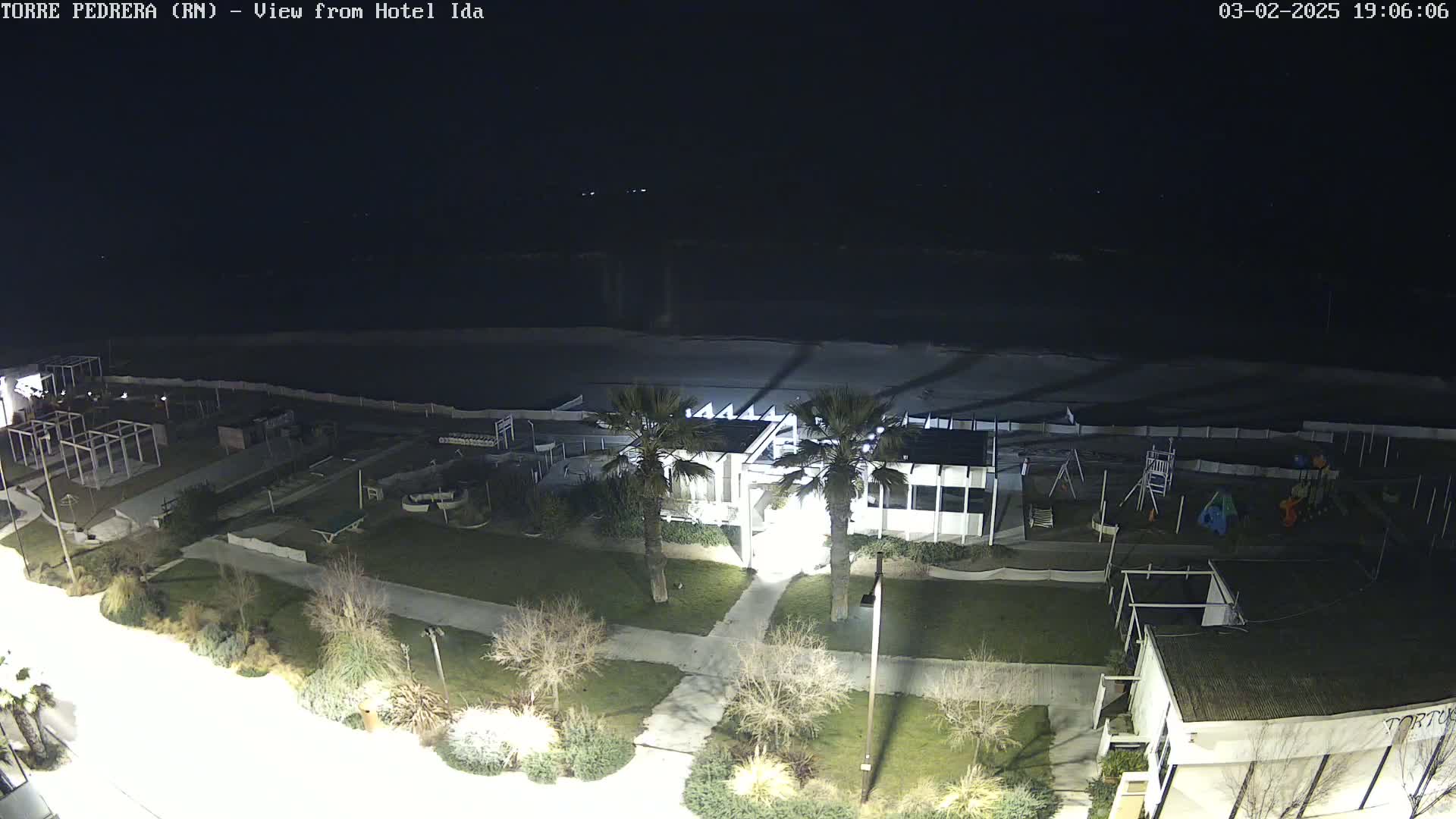 A nighttime, overhead view of a beachside area shows a mostly empty playground, a building, palm trees, and a dark, wavy ocean under a clear night sky.