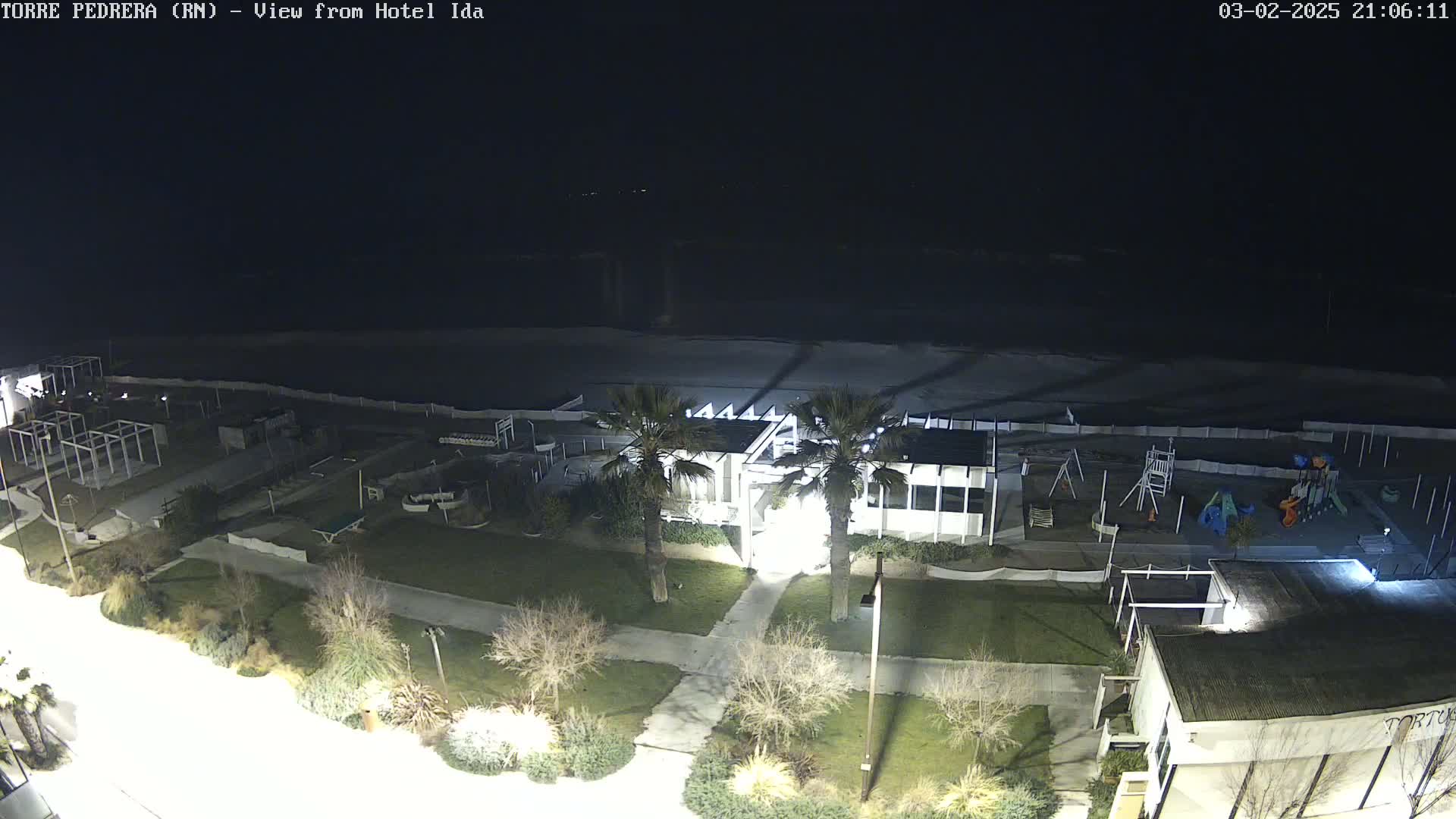 A nighttime aerial view of a beach area shows a building, playground, and palm trees illuminated under dark skies.