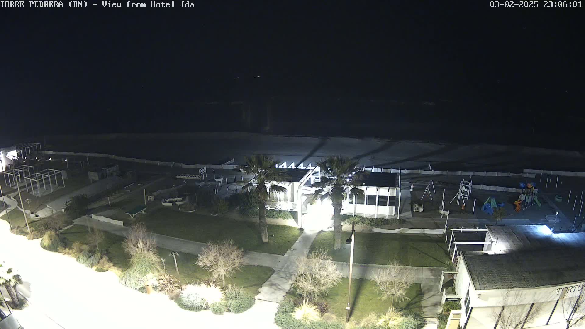 A nighttime, aerial view of a beachside area shows a building with palm trees, a playground, and a mostly empty beach under a dark sky.