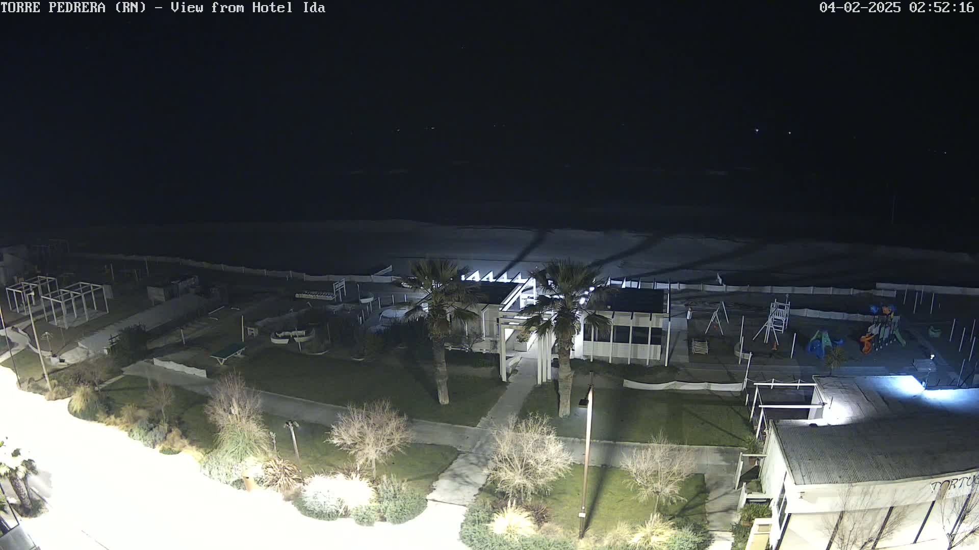 A nighttime aerial view shows a beach, a playground, some buildings, and palm trees under a dark, clear sky.