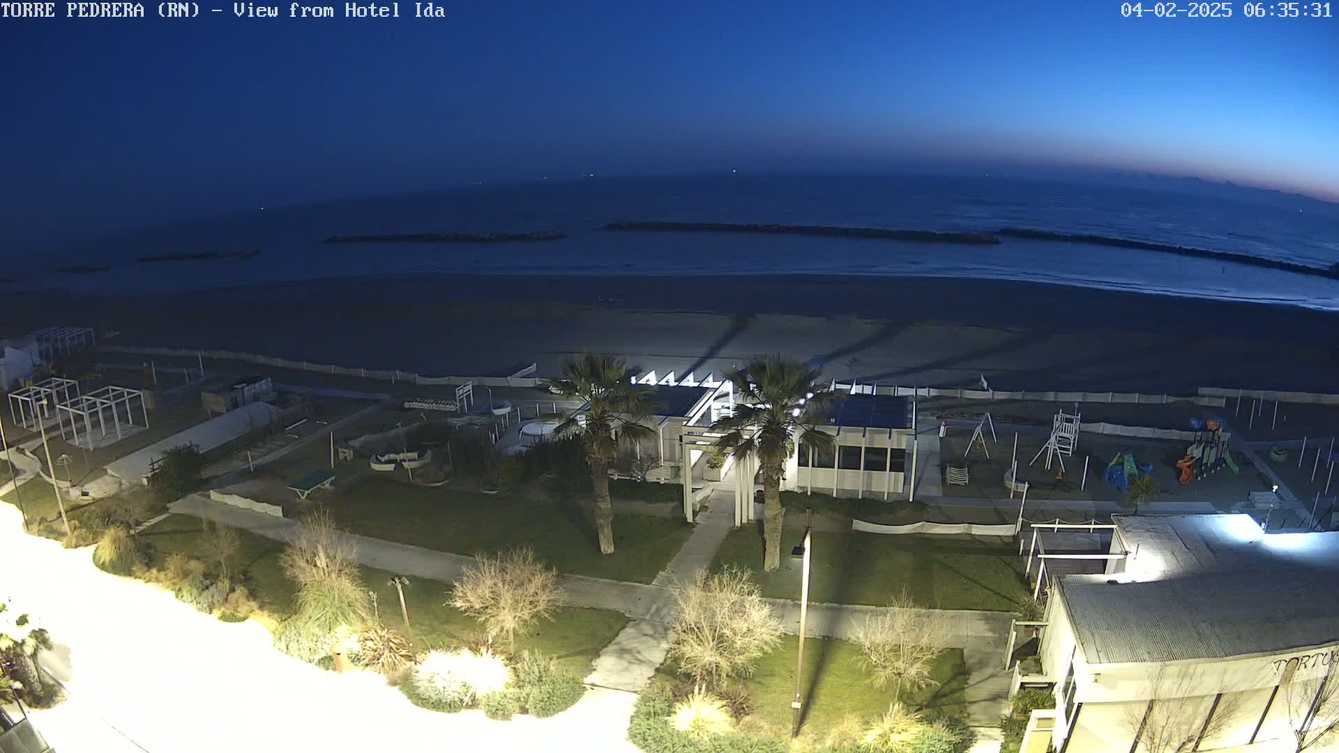 A high-angle, nighttime view shows a beach, a playground, and a building near the ocean under a dark blue, twilight sky.