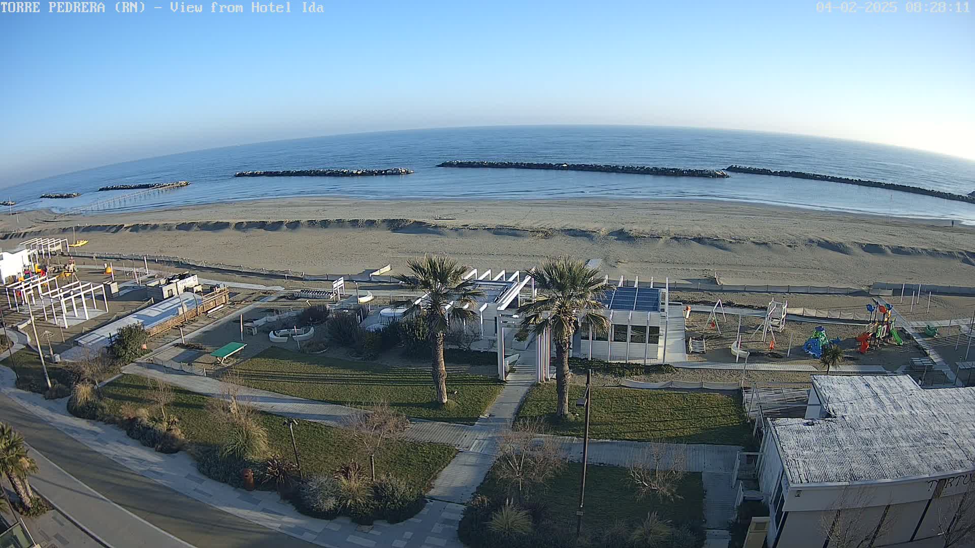 A sandy beach with calm ocean waves under a clear blue sky, featuring a beachfront playground, palm trees, and a modern building.